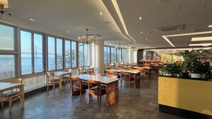Ein geräumiges Restaurant mit großen Fenstern und Blick auf eine Brücke und Wasser, Holztische und -stühle sowie eine Theke mit Pflanzen.