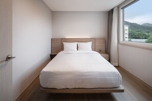 A neatly made bed in a hotel room with a wooden headboard, a nightstand, and a window with a view of greenery and buildings. - Kumho Sulak Resort (Sokcho)