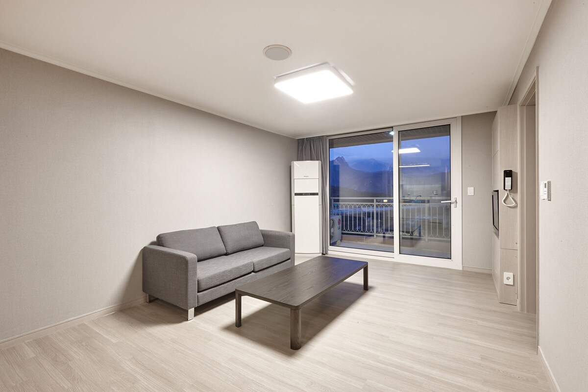 Un salon moderne avec un canapé gris, une table basse en bois et une grande porte-fenêtre coulissante donnant sur un balcon.