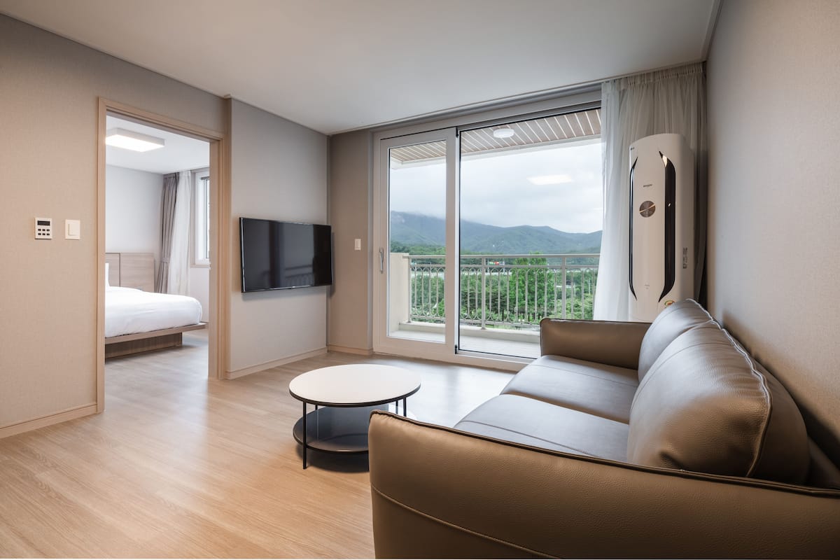 Habitación de hotel moderna con sofá, una mesita redonda, televisor y balcón con vistas a montañas.