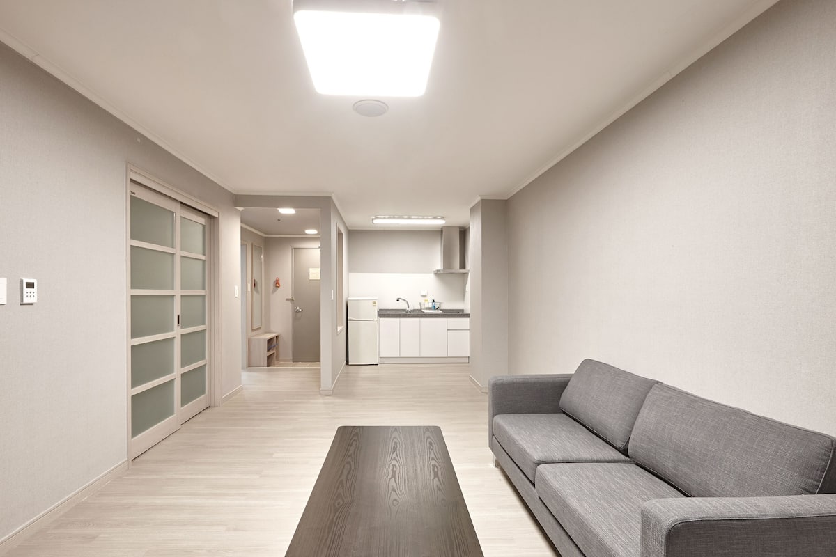 Una sala moderna con un sofá gris, una mesa de centro de madera y una cocina al fondo.