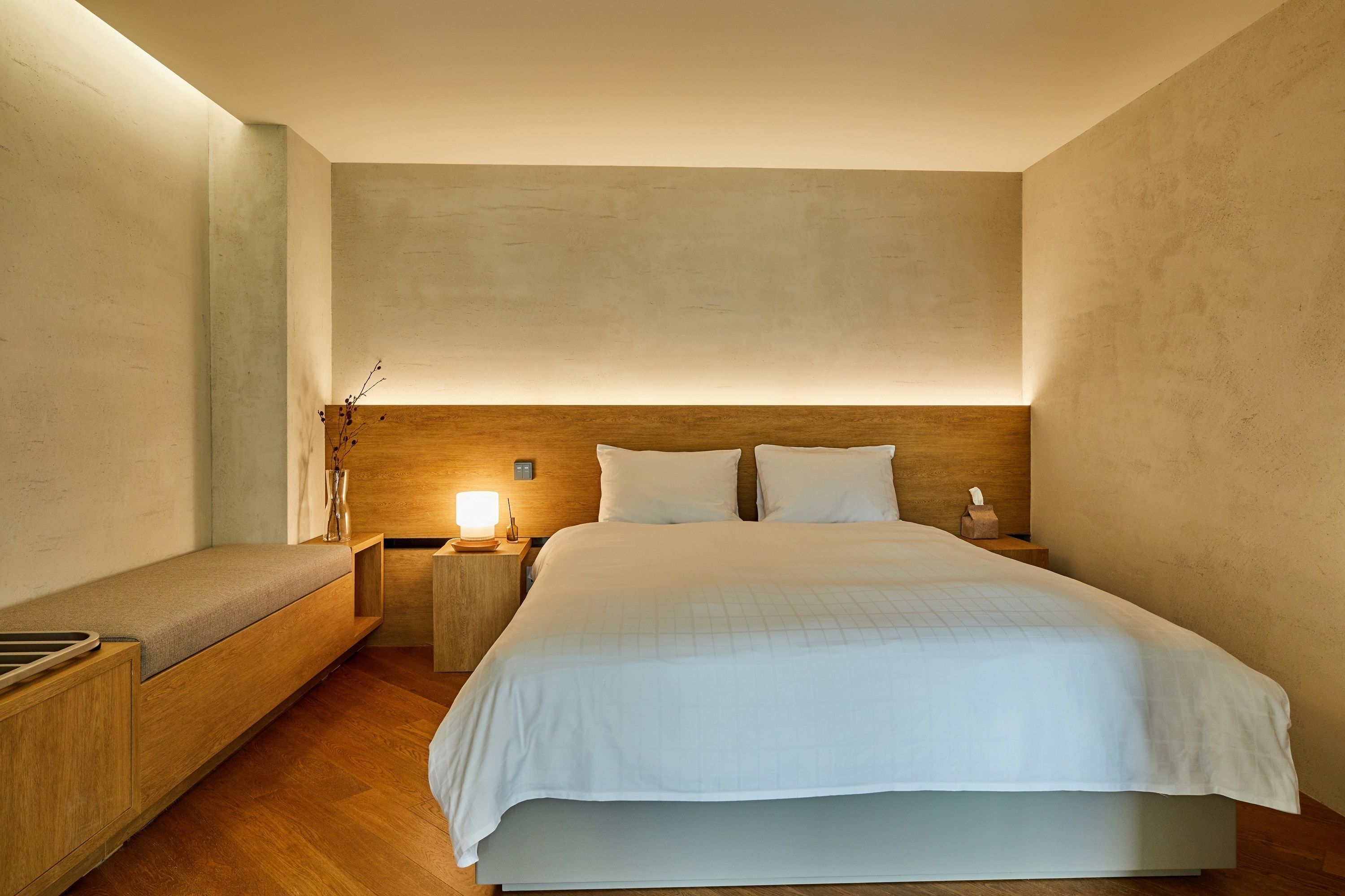 A modern hotel room with a large bed, a wooden bench, a nightstand, and a bedside table.