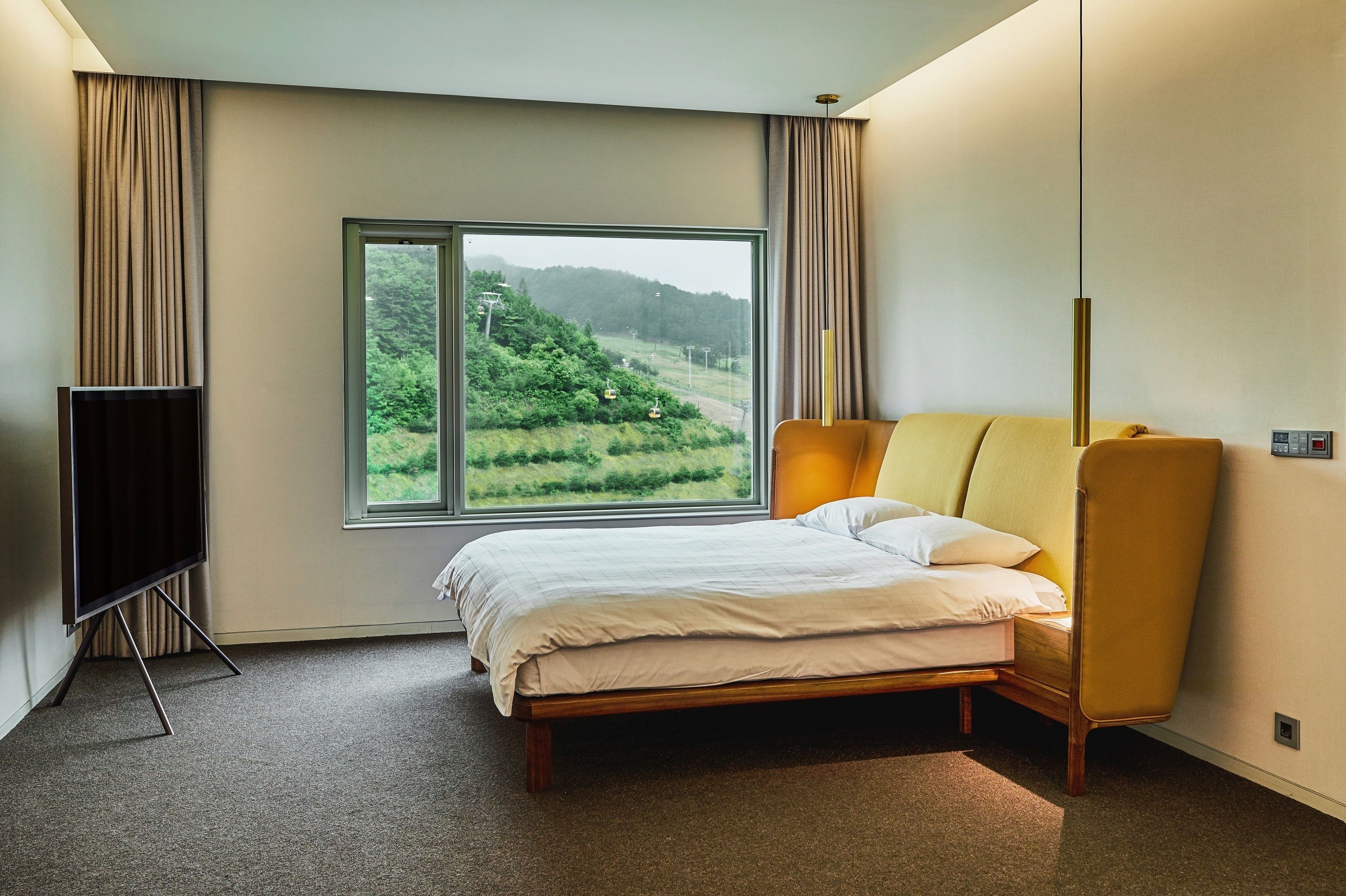 Une chambre avec une grande fenêtre, un lit, un téléviseur posé sur un meuble et une lampe moderne.