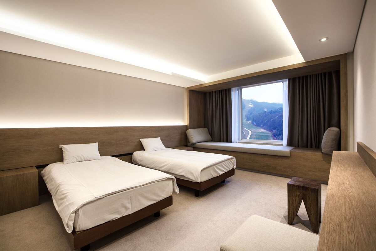 Una habitación de hotel moderna con dos camas, un banco, una mesita pequeña y un ventanal con vistas.
