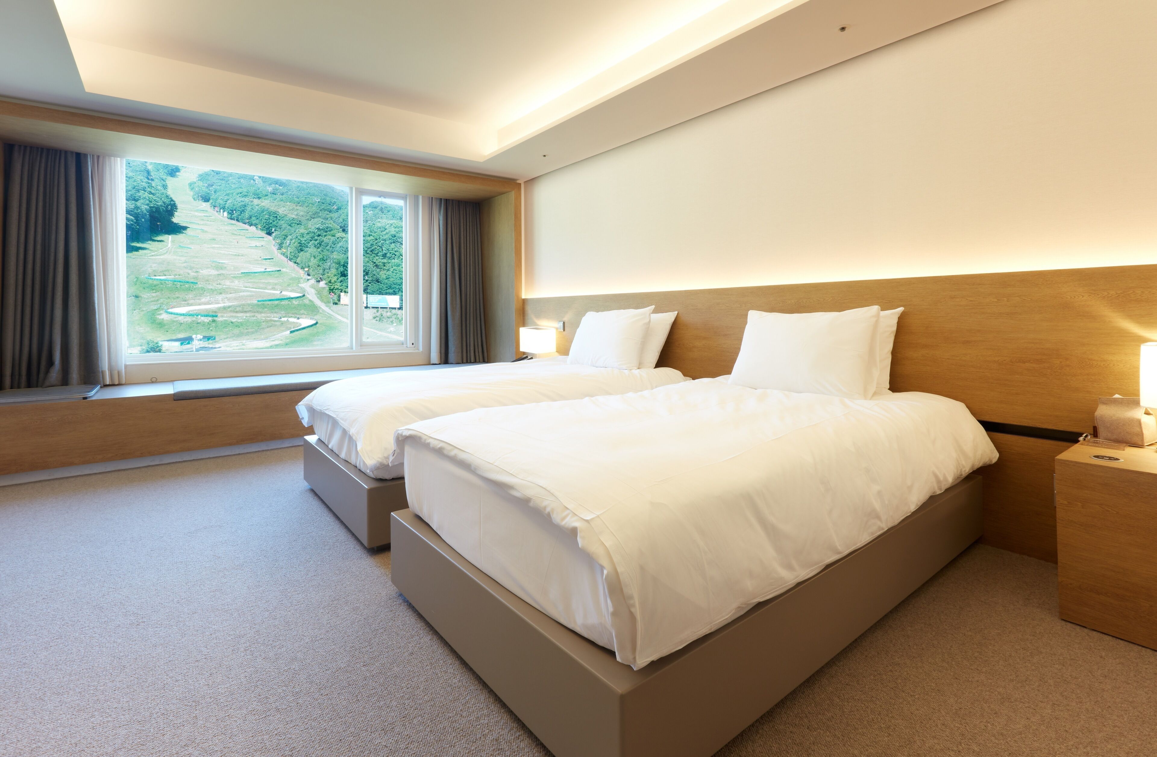Habitación de hotel con dos camas, un ventanal grande que ofrece vistas a un área boscosa y un cabecero de madera.