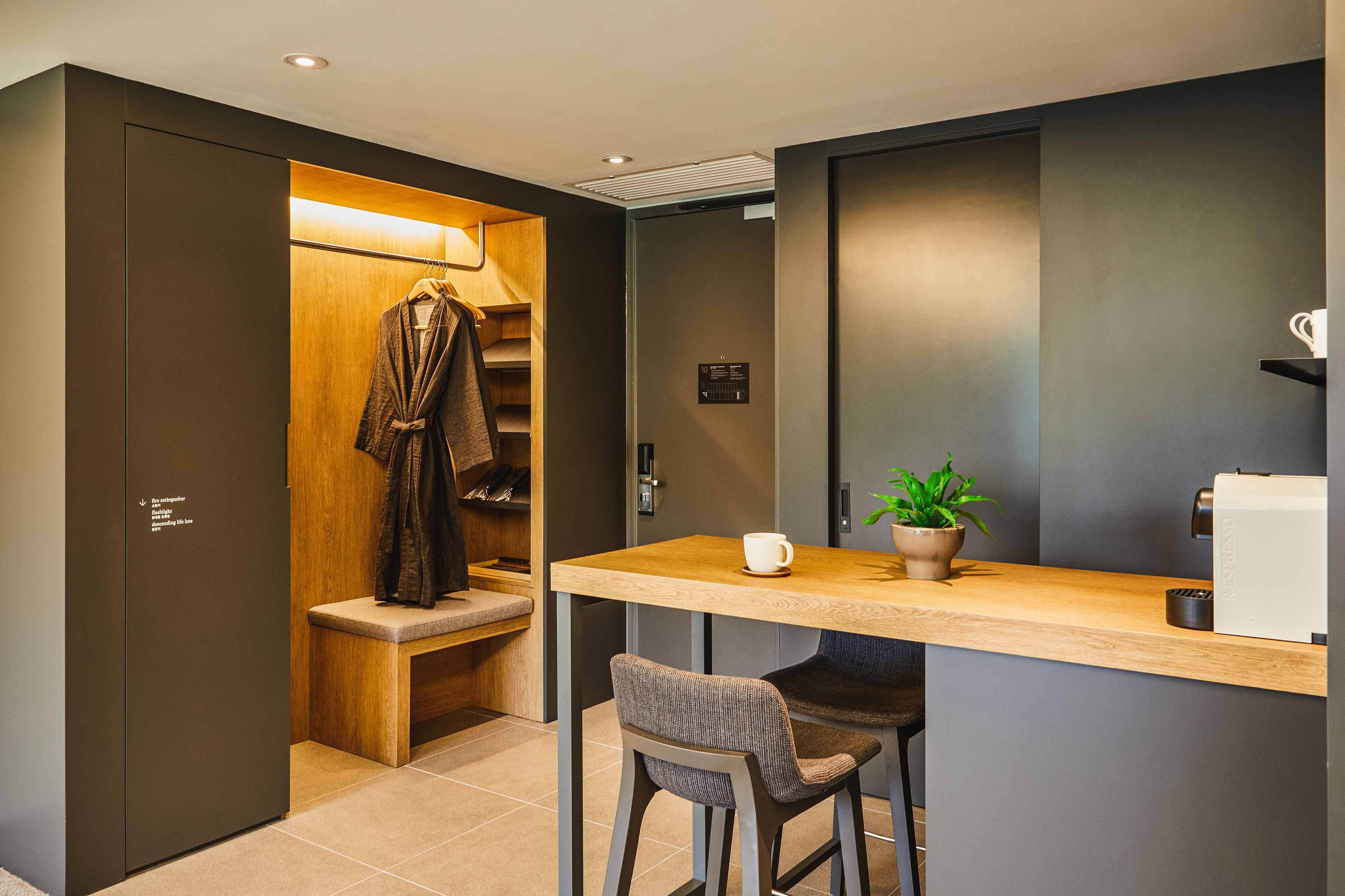 A modern hotel room with a wooden closet, a small seating area with a bench and chairs, a coffee machine, and a potted plant.