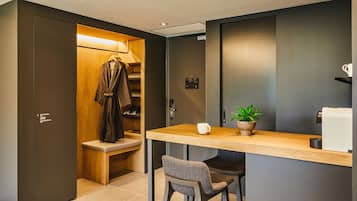A modern hotel room with a wooden closet, a small seating area with a bench and chairs, a coffee machine, and a potted plant.