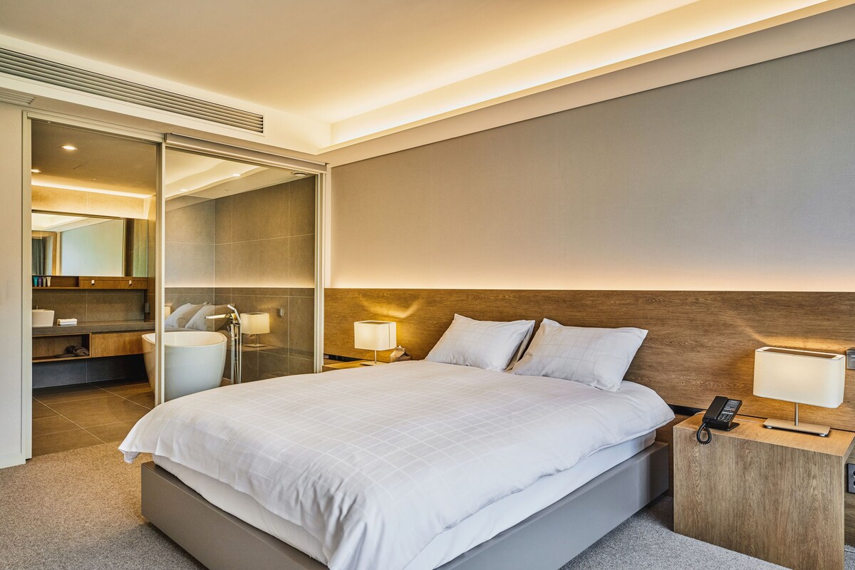 Una habitación de hotel moderna con una cama grande, lámparas de noche, un cabecero de madera y un box de ducha de vidrio.