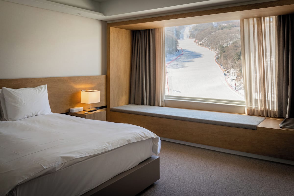 Hotellihuone, jossa on suuri sänky, yöpöytä lampun kanssa, ikkunasyvennys verhoilla sekä näkymä lumiseen maisemaan.