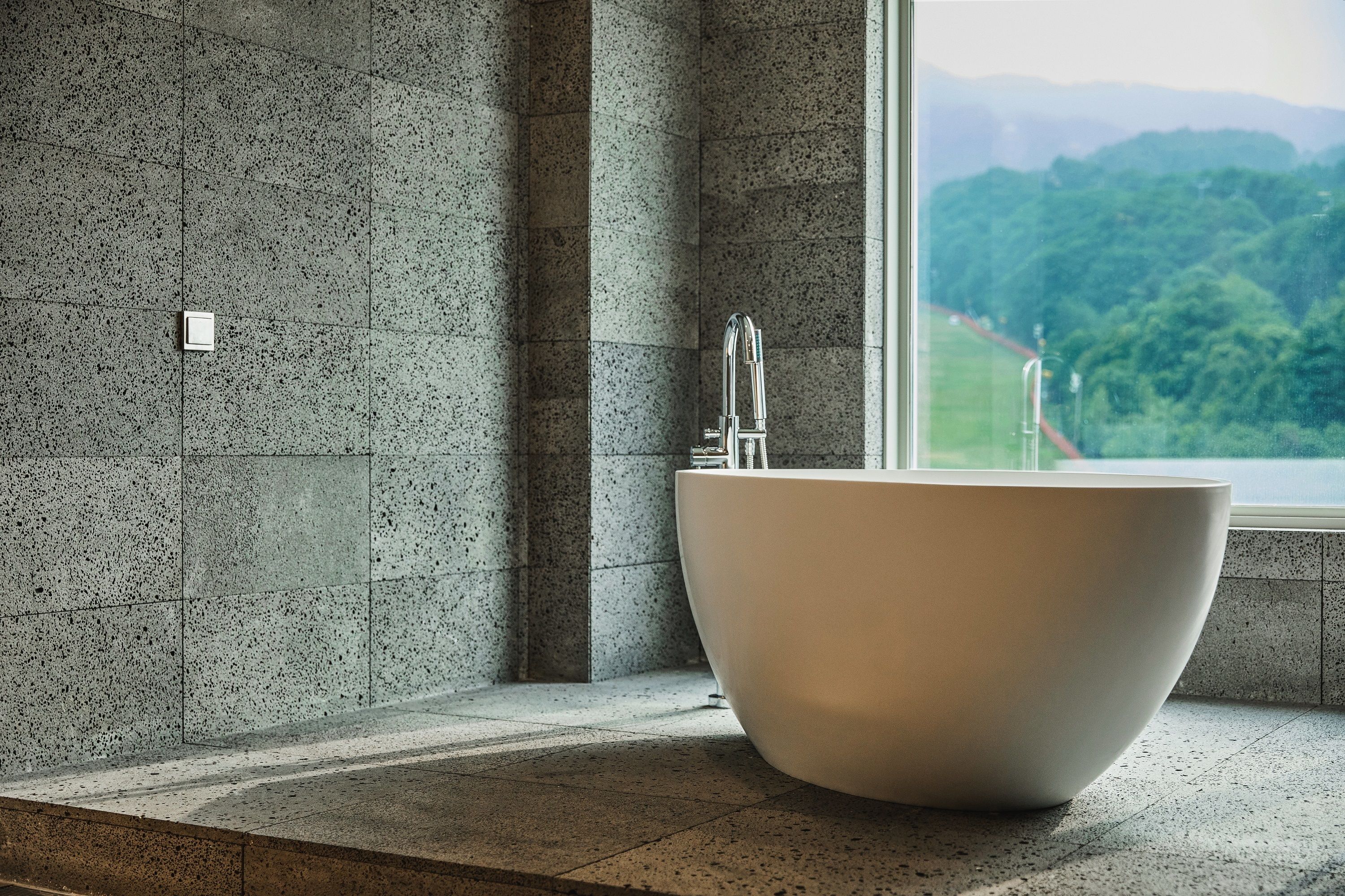 A modern bathroom with a large freestanding bathtub, a visible window with a view of greenery, and a tiled wall.