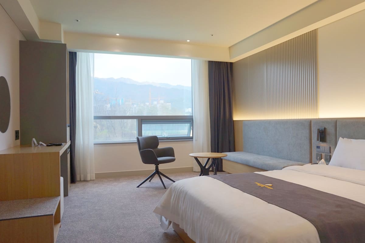 A hotel room with a bed, a desk, a chair, a small table, and a large window with a city view.