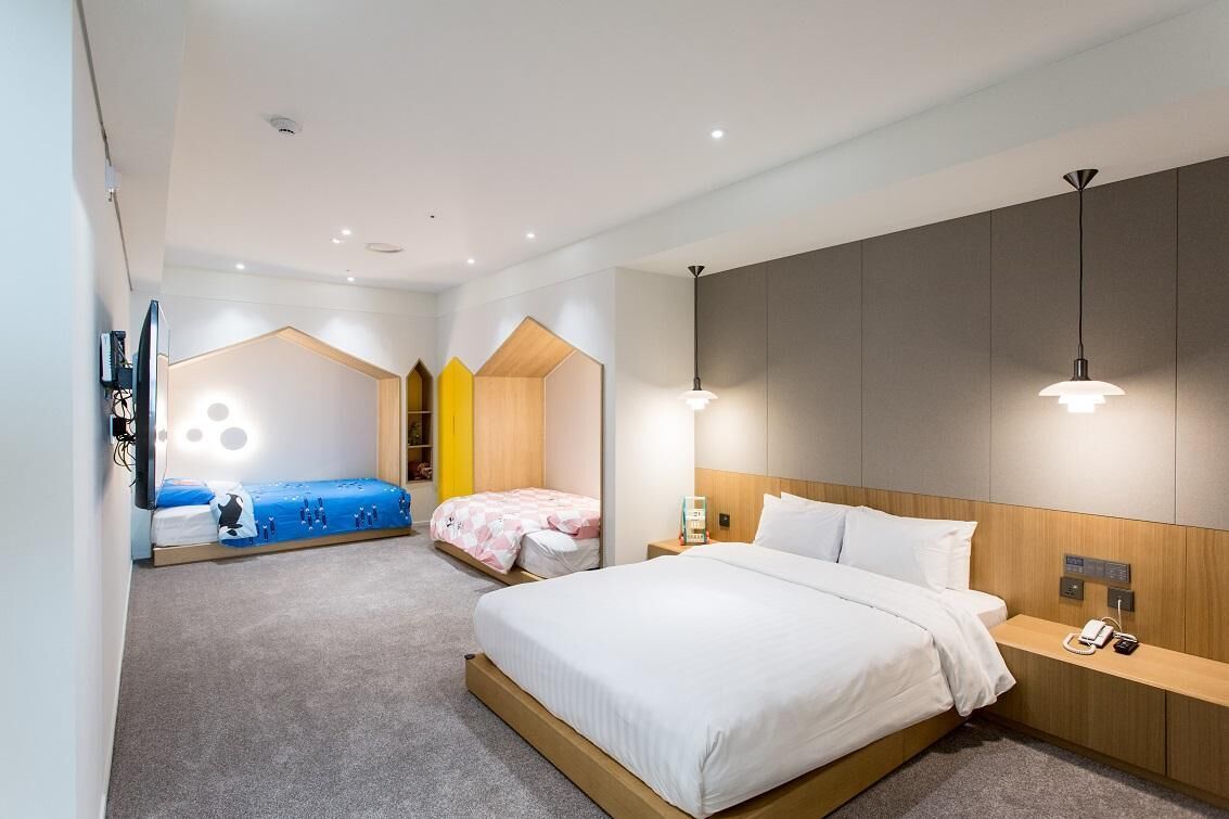 A modern hotel room with a large bed, a desk, and a TV. There are two smaller beds in the background.