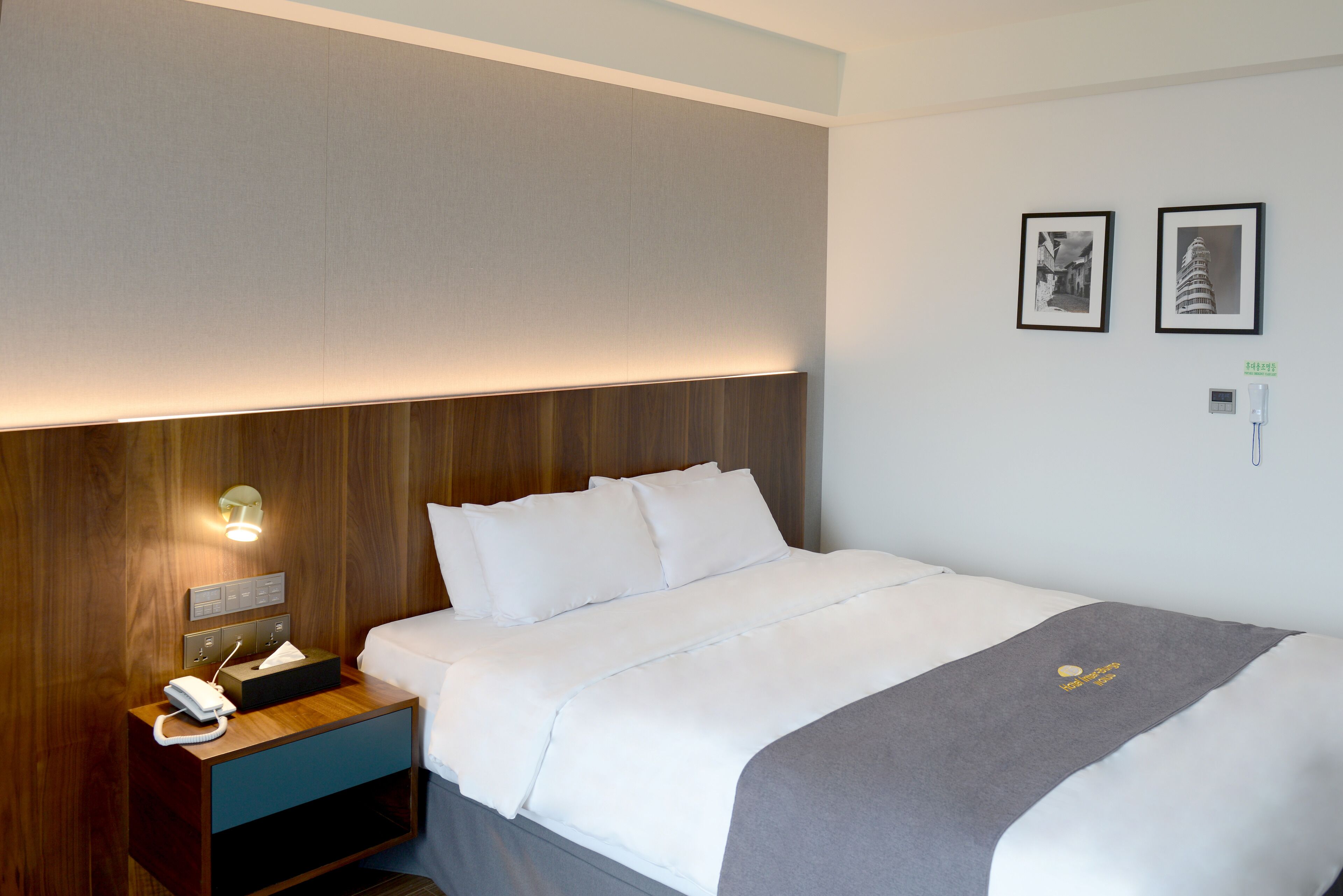 Una habitación de hotel con una cama bien hecha, una mesita de noche con lámpara y dos cuadros en la pared.