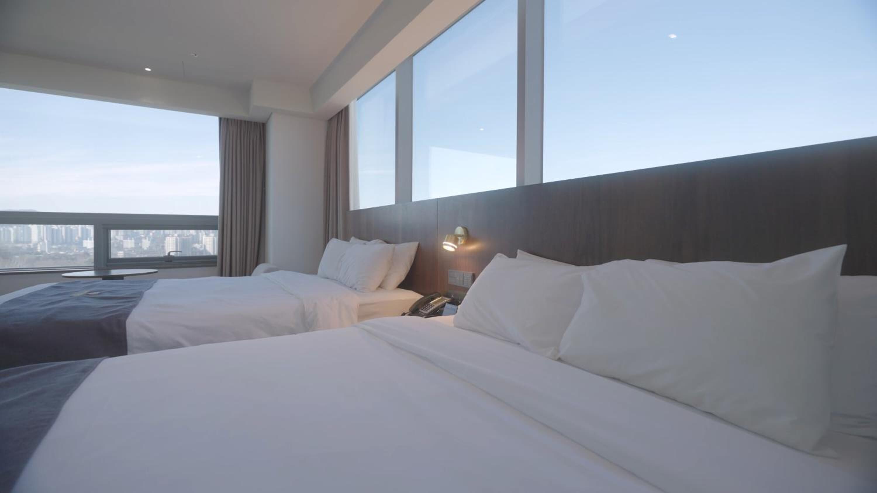 A hotel room with two beds, a bedside table with a lamp, and a view of the cityscape through large windows.