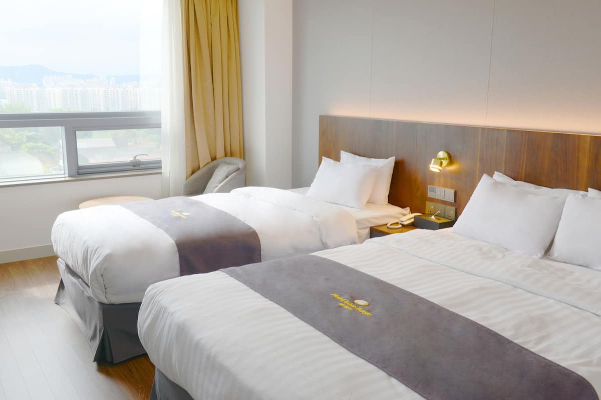 A hotel room with two beds, a window with curtains, a bedside table with a lamp, and a view of the cityscape.