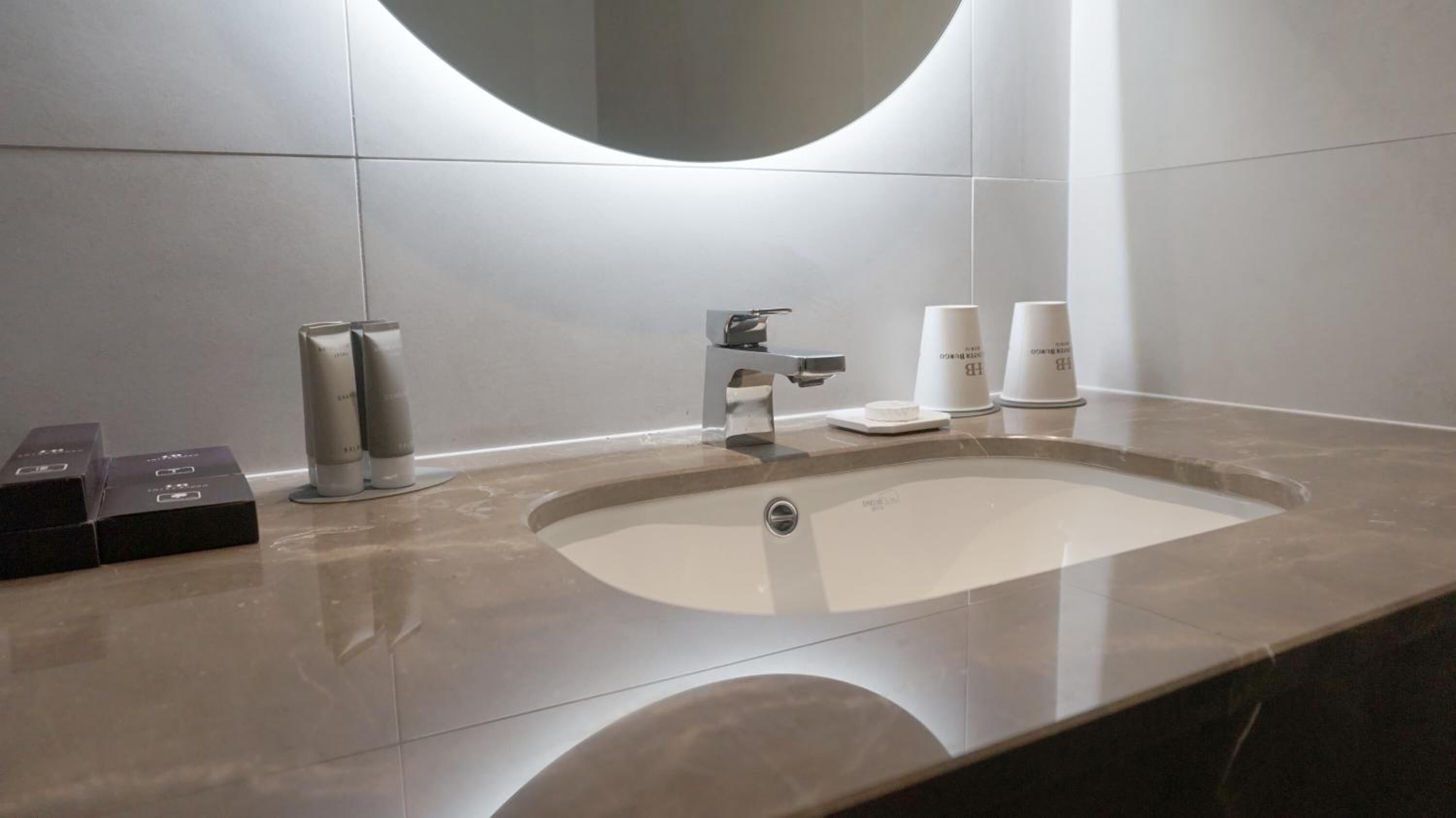 A modern bathroom with a large sink, a mirror, and a faucet. There are two white cups and a box on the counter.