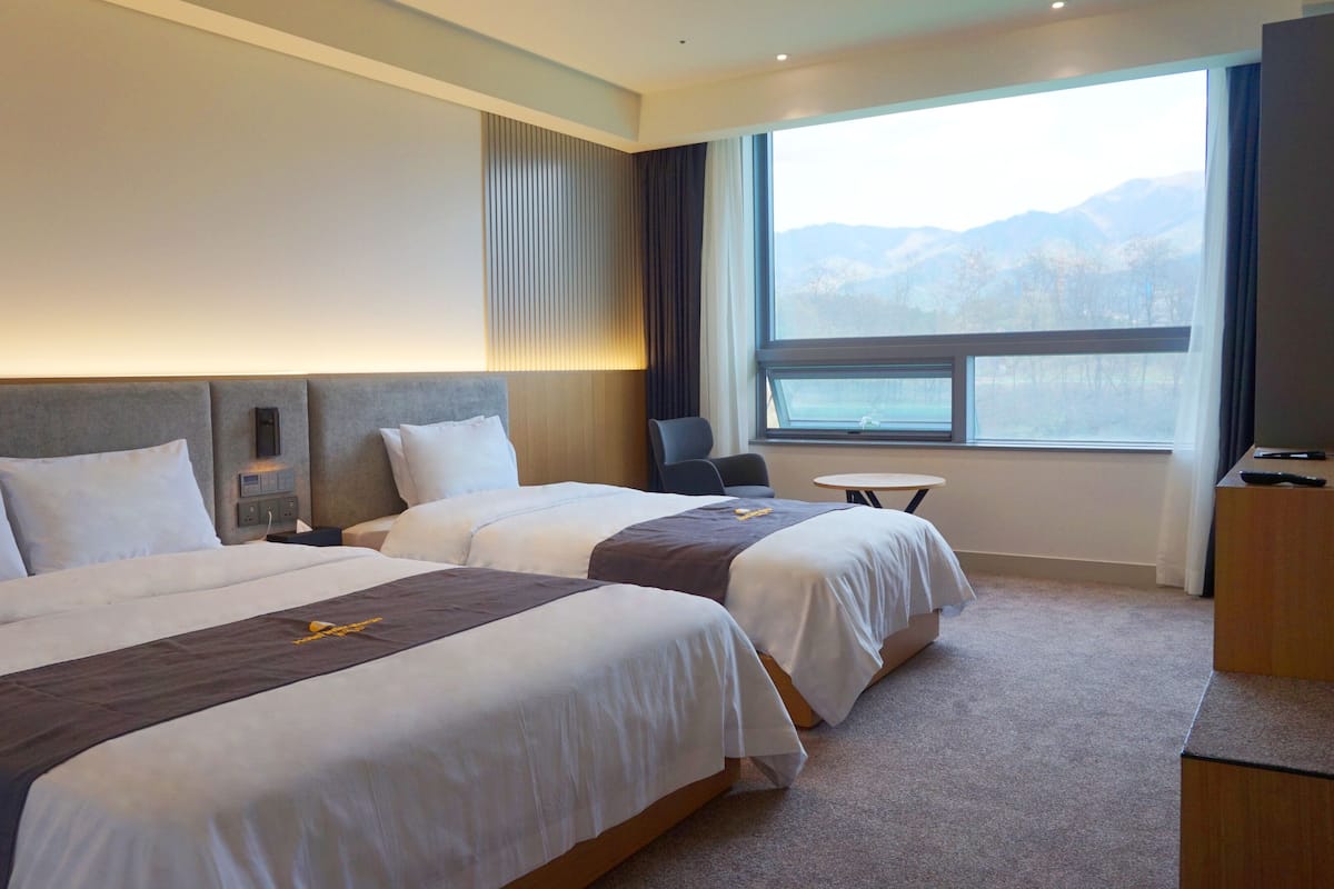 A hotel room with two beds, a small table, a desk, and a large window with a mountain view.