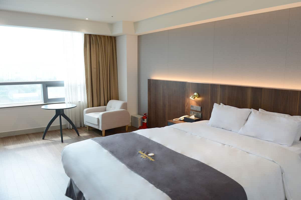A modern hotel room with a large bed, a small table, a chair, and a view of the city through the window.