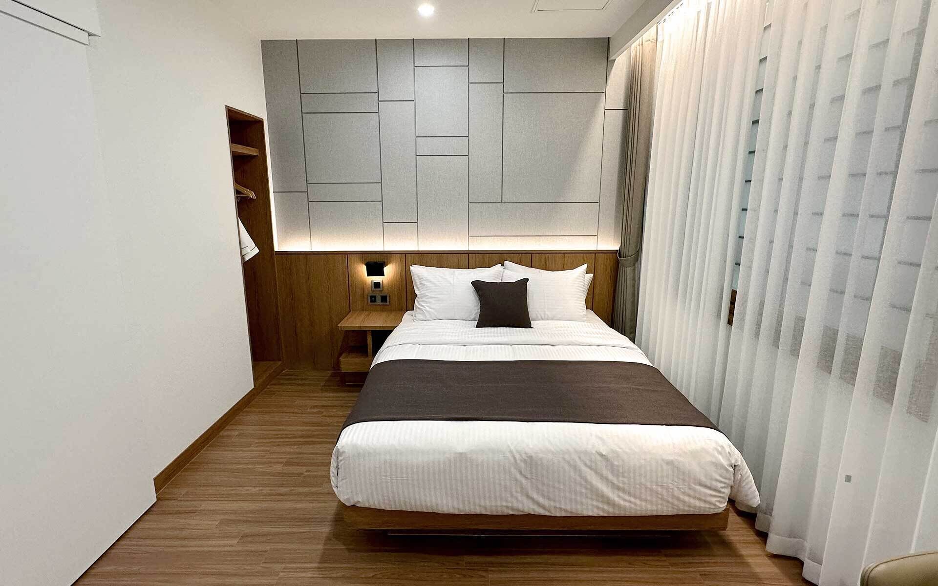 A modern hotel room with a large bed, wooden flooring, a geometric wall design, and a window with curtains.