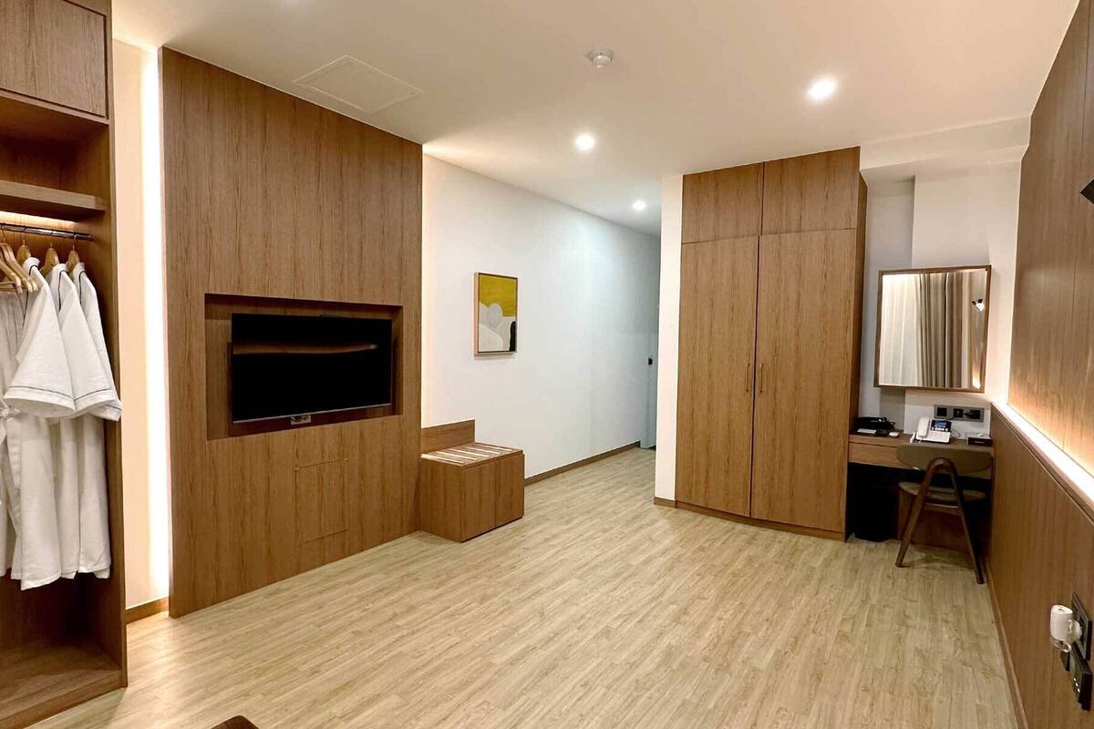 A modern hotel room with wooden paneling, a flat-screen TV, a desk with a chair, a wardrobe, and a bathroom mirror.