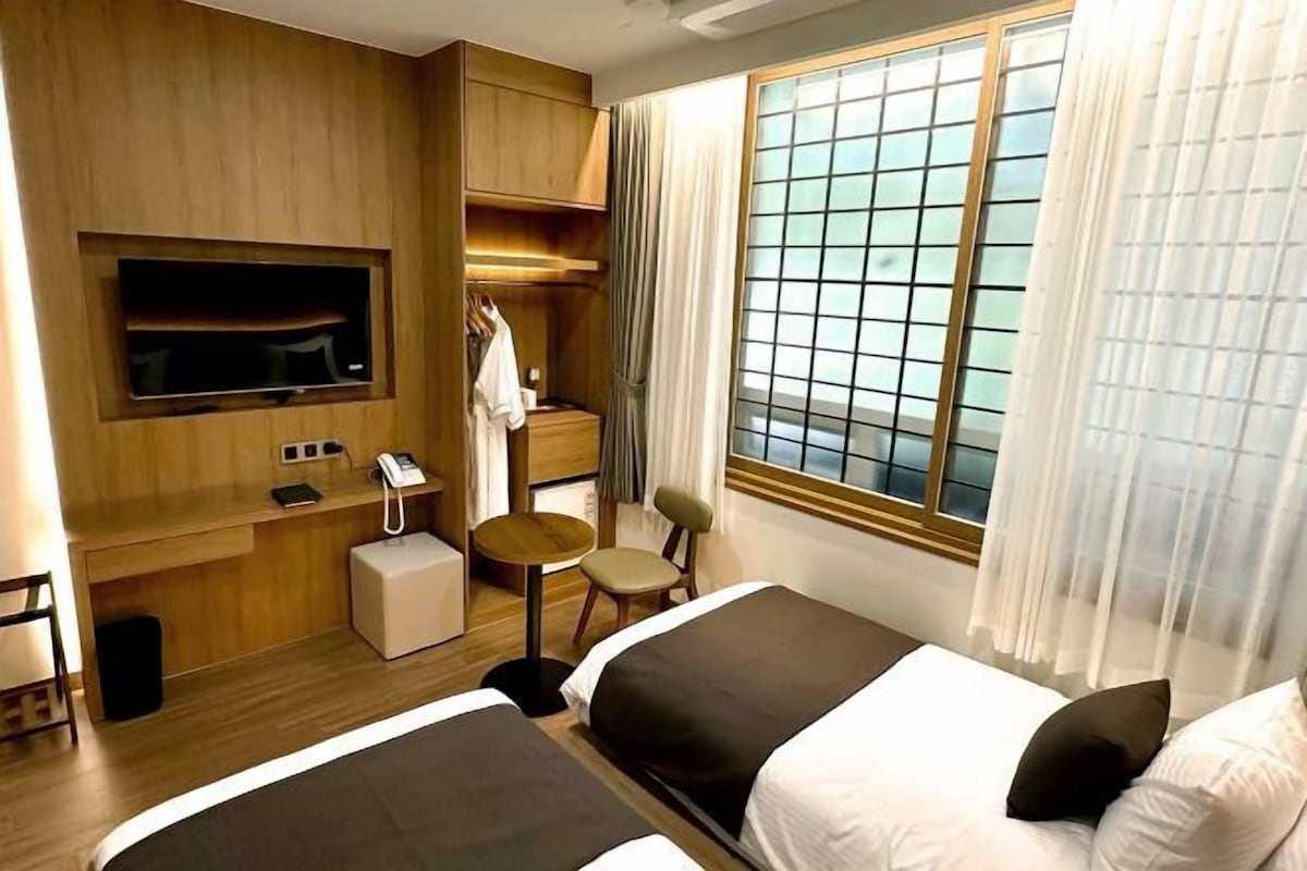 A hotel room with two beds, a TV mounted on the wall, a desk with a phone, a small table, and a window with curtains.