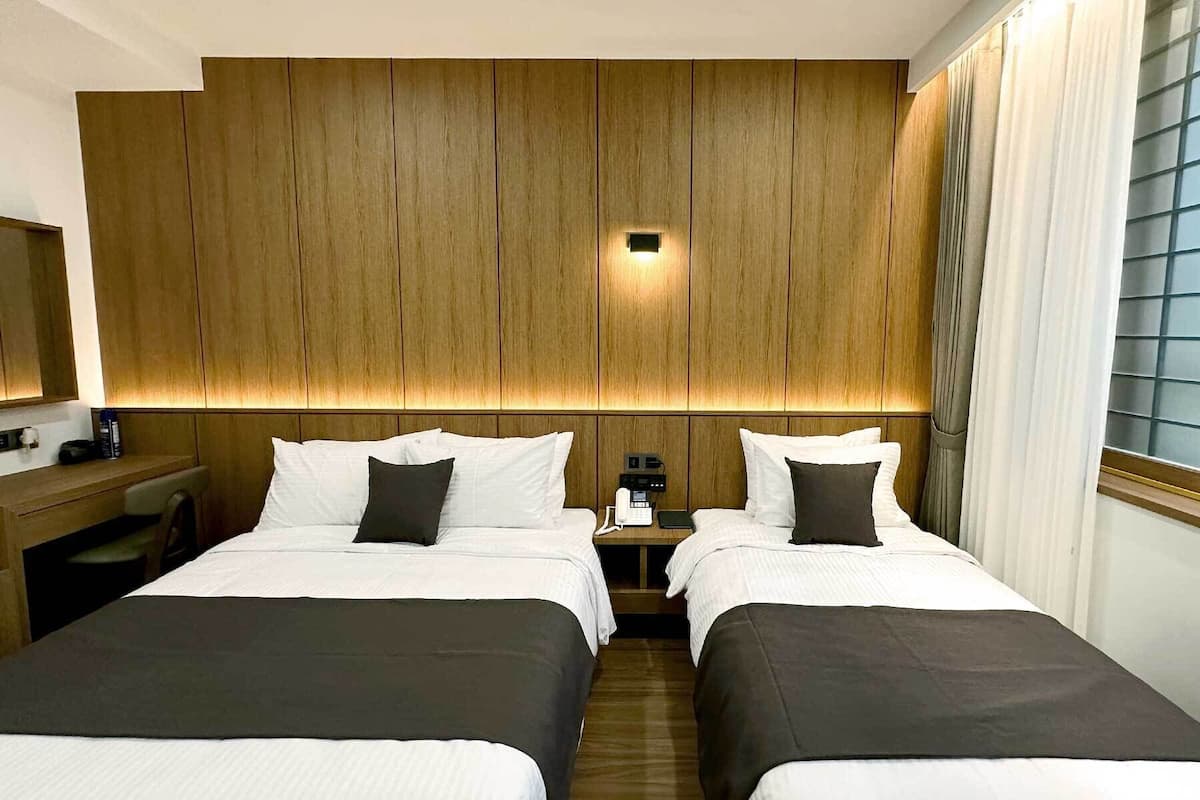 A hotel room with two beds, wooden paneling on the wall, a bedside table with a phone, and a window with curtains.
