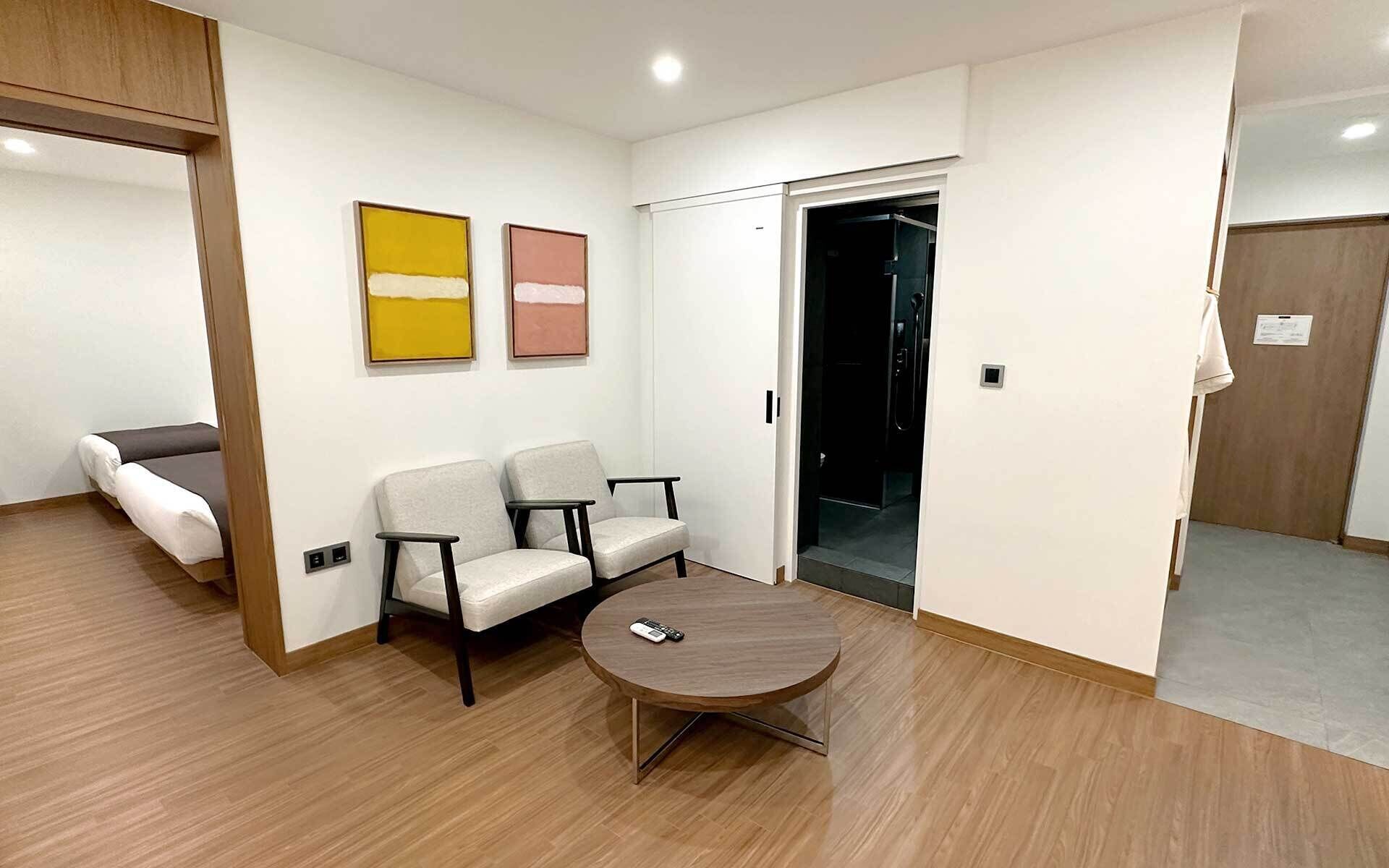 Une chambre d’hôtel moderne avec un plancher en bois, une table ronde, deux fauteuils, un canapé, une porte coulissante et deux tableaux abstraits accrochés au mur.