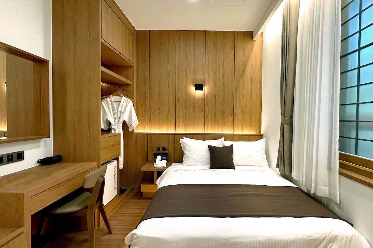A hotel room with a bed, a desk, a chair, a wardrobe, and a bedside table with a lamp.