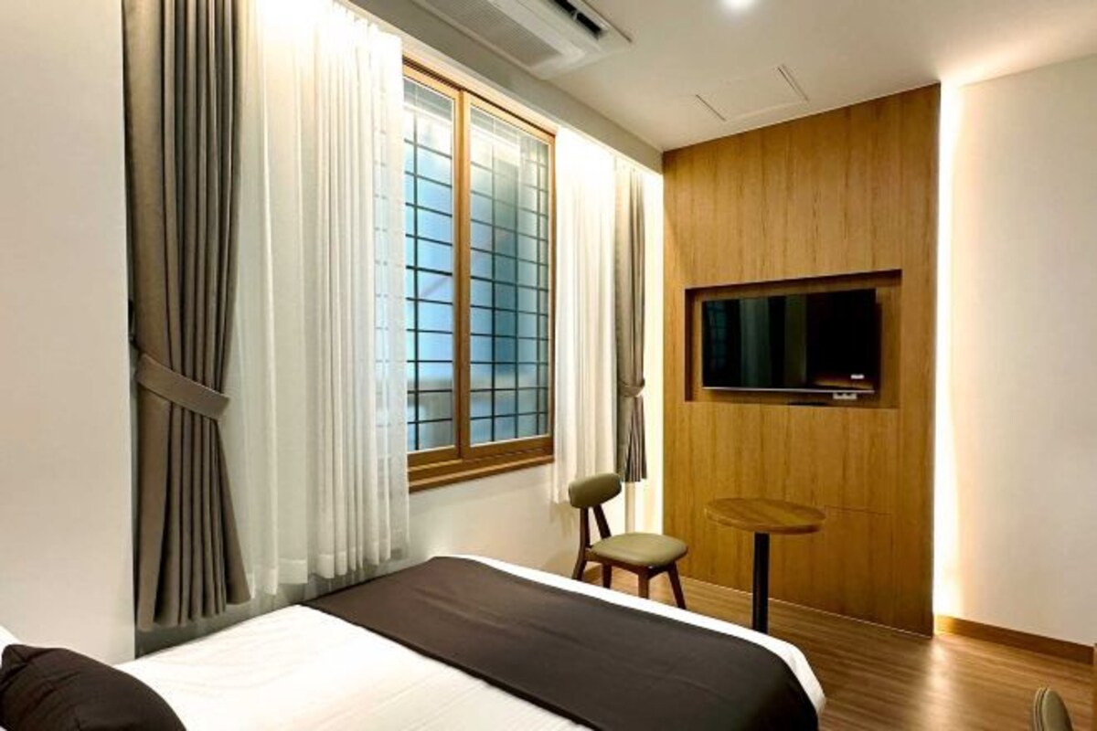 Quarto de hotel moderno com uma cama grande, uma mesinha, uma cadeira e uma televisão fixada na parede.