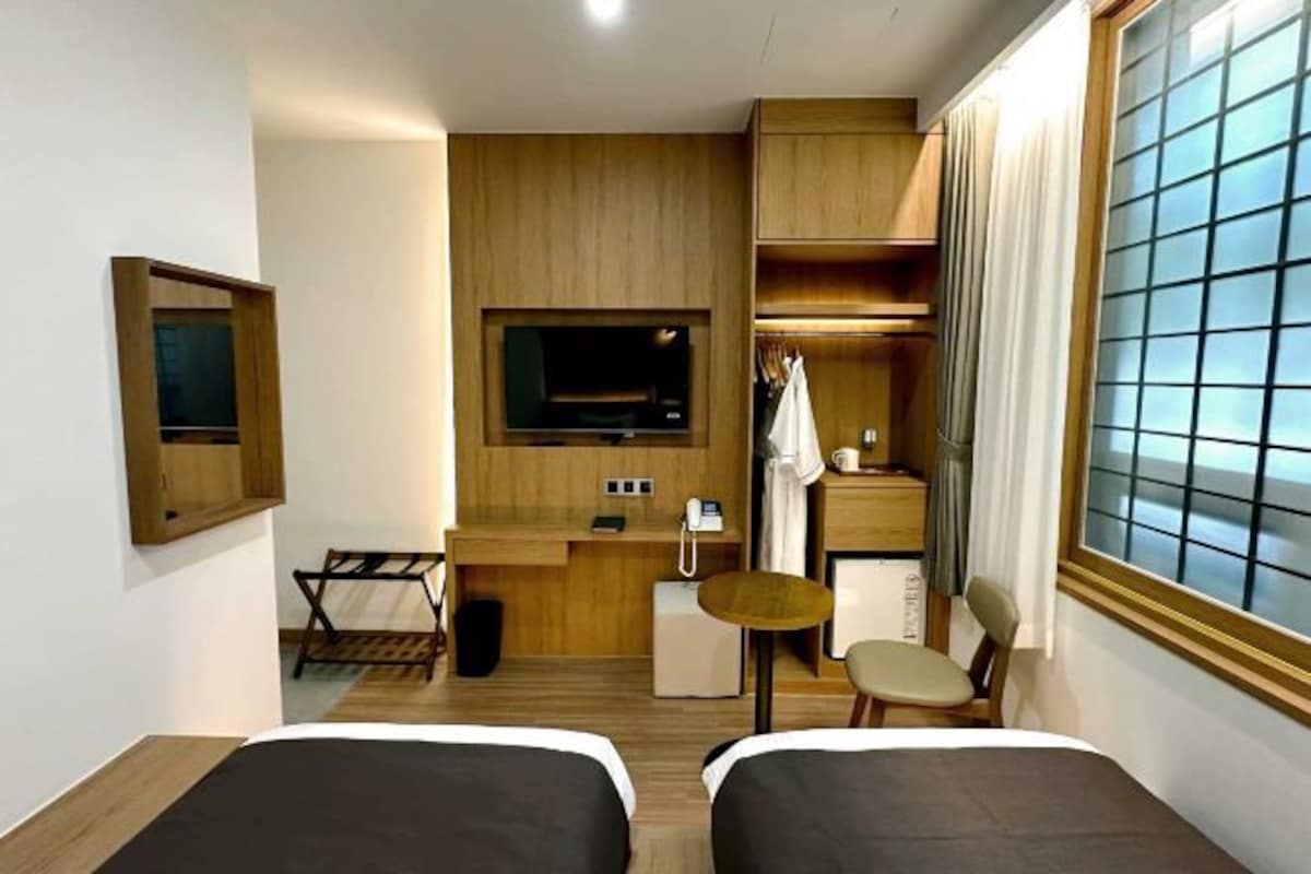 Quarto de hotel compacto com duas camas, uma escrivaninha, uma cadeira, uma mesinha, televisão e janela com cortinas.