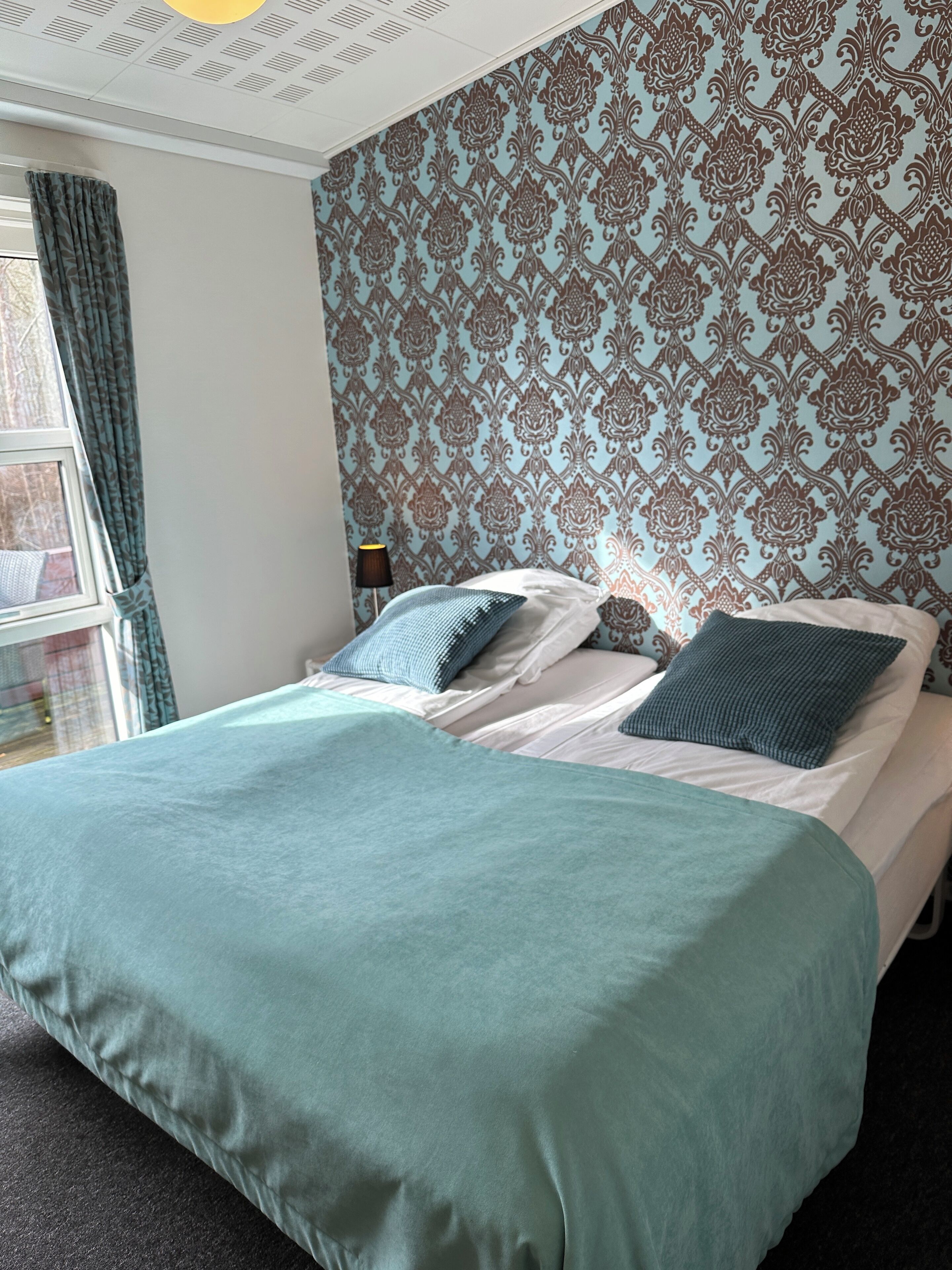 Een hotelkamer met een bed, een raam met gordijnen en een behang met een patroon.