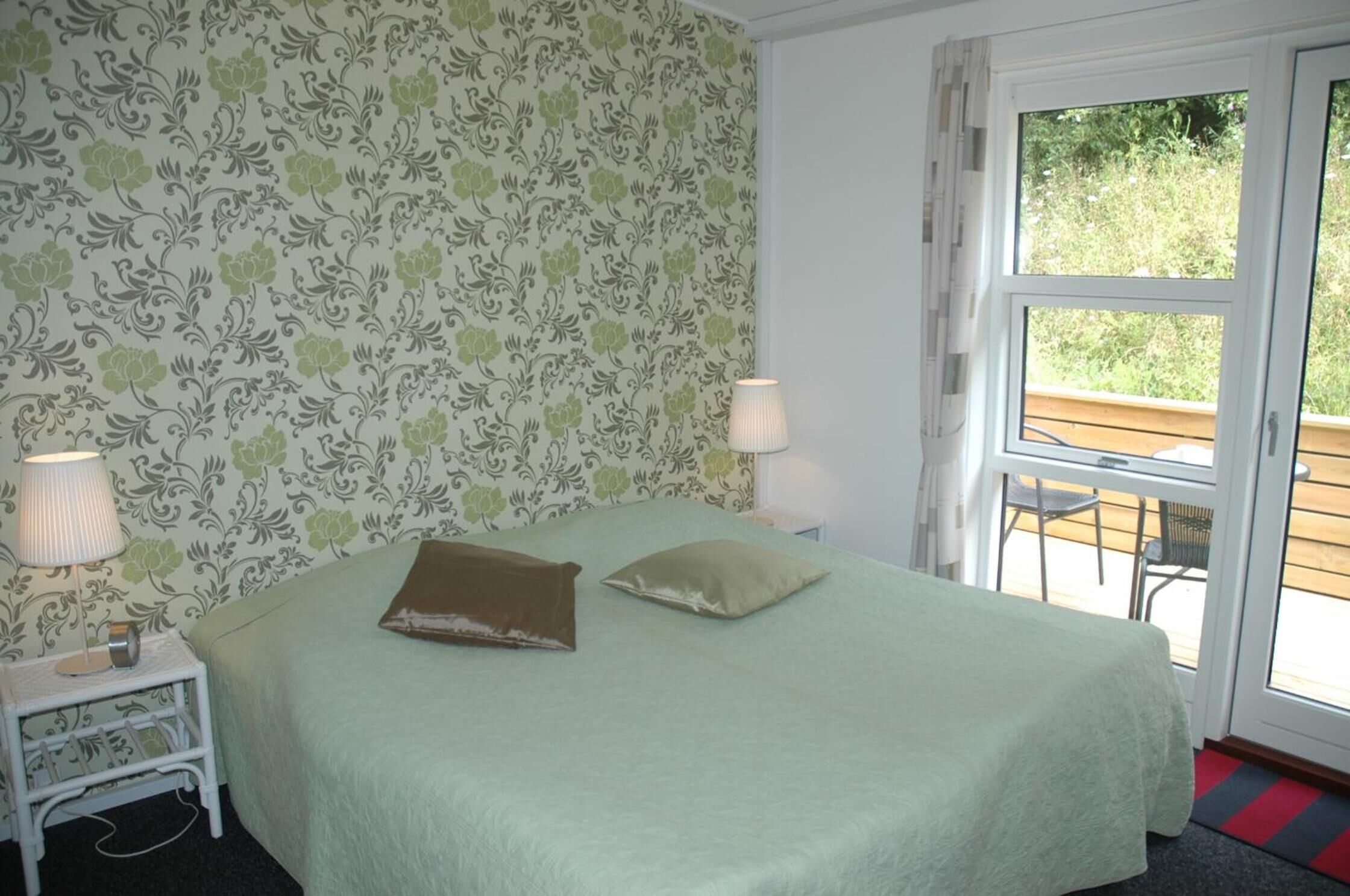 A room with a bed, two bedside lamps, a patterned wallpaper, and a view through a glass door to an outdoor area.