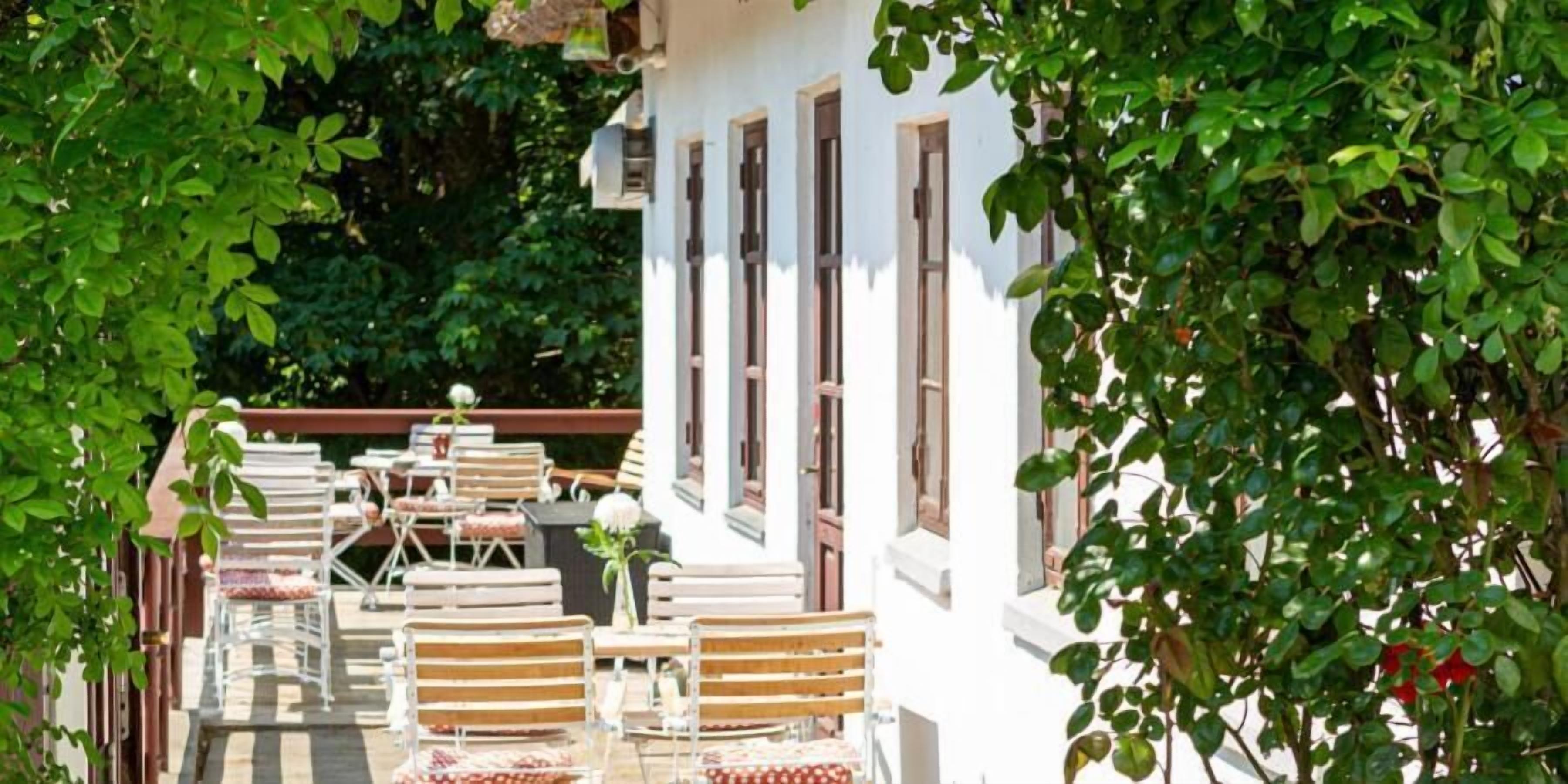 Un área de estar al aire libre con mesas y sillas, rodeada de follaje y un edificio blanco con ventanas.