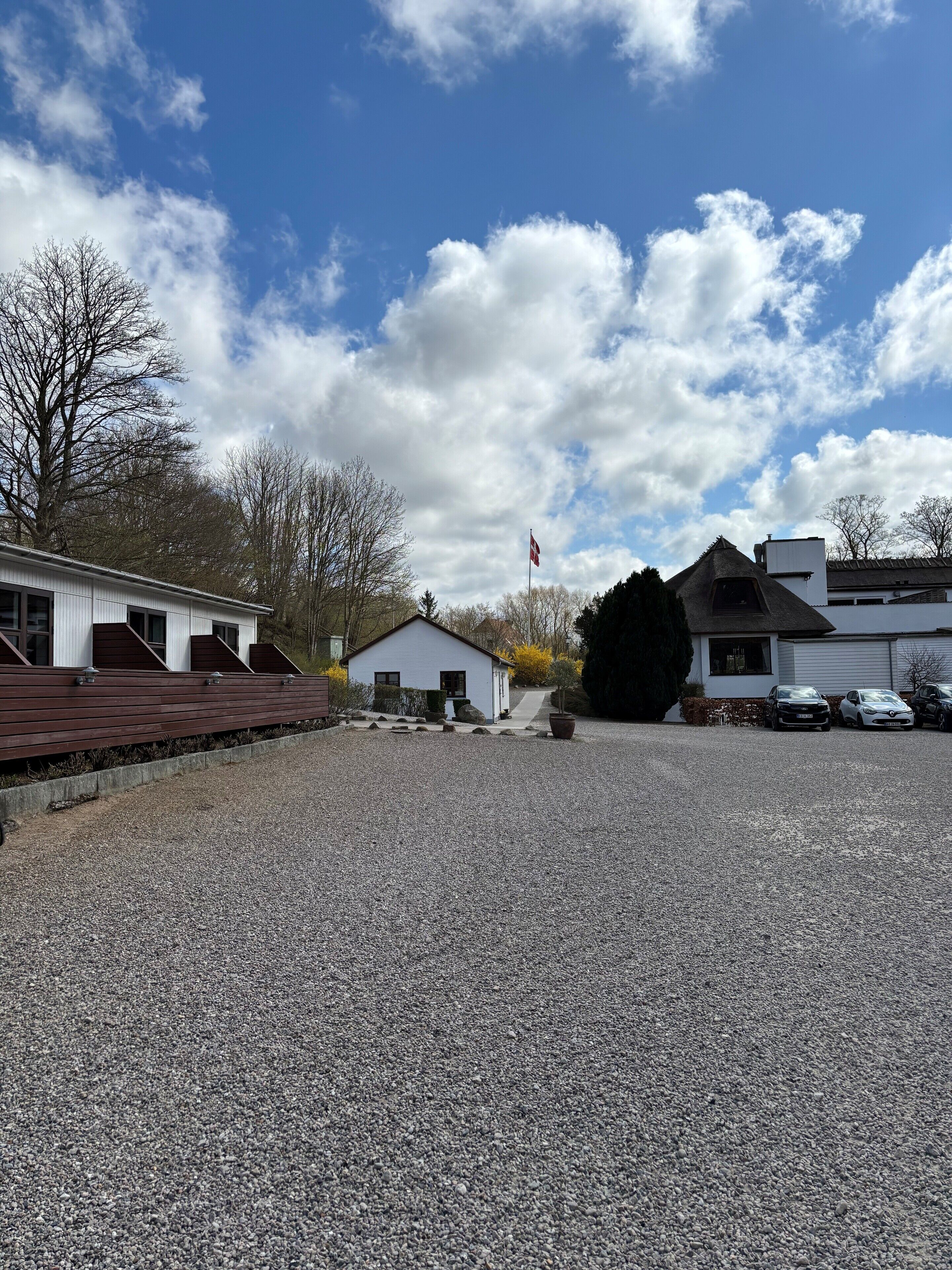 A gravel parking area with a white building, a flag, and a row of parked cars.