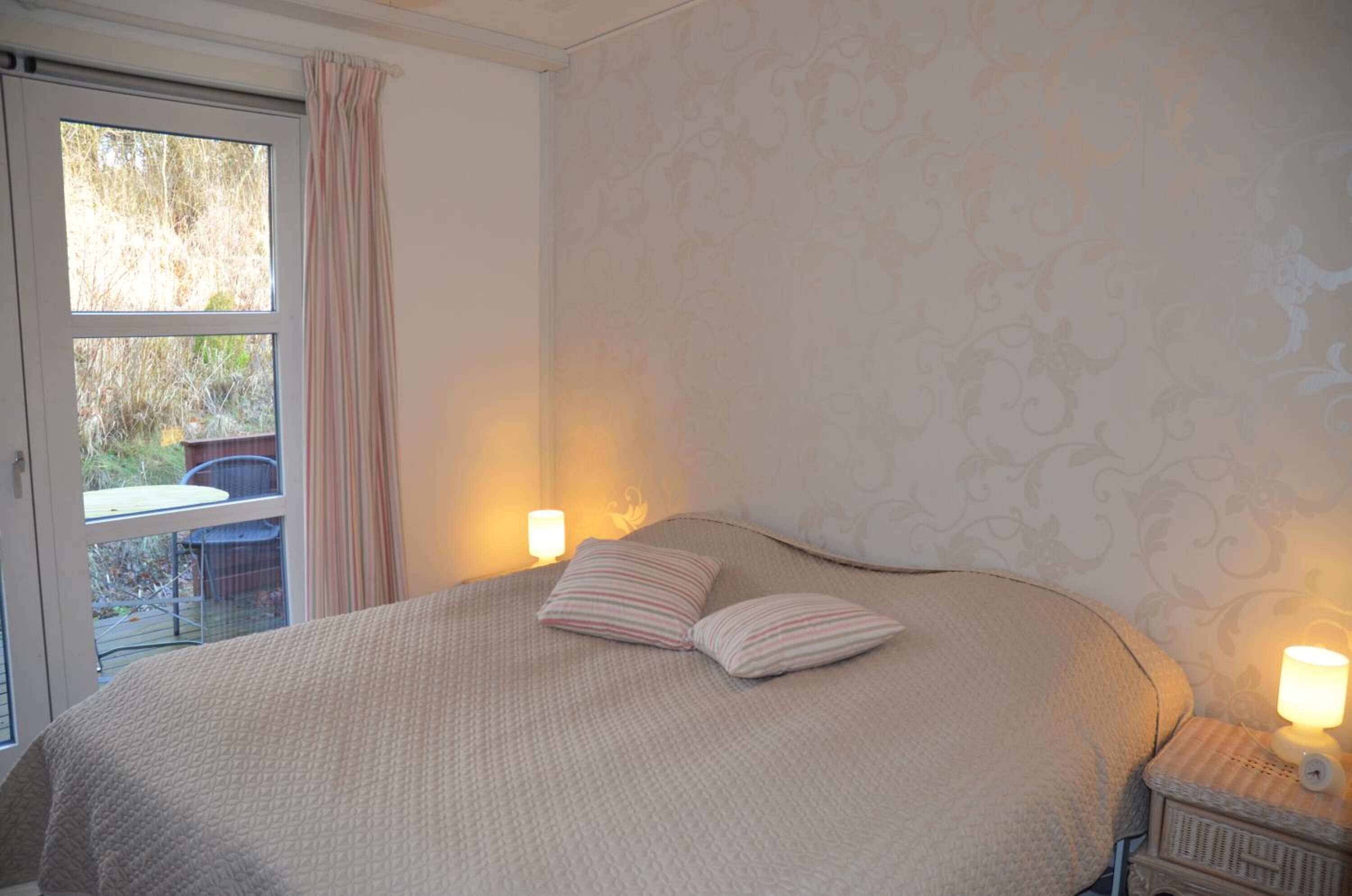 Quarto com cama, duas luminárias de cabeceira, janela com cortinas e vista para o exterior.