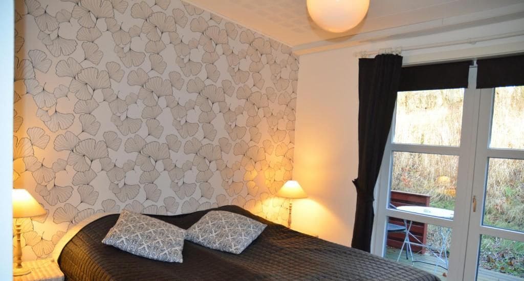 A bedroom with a patterned wallpaper, a bed with a dark quilt, two pillows, bedside lamps, and a window with curtains.