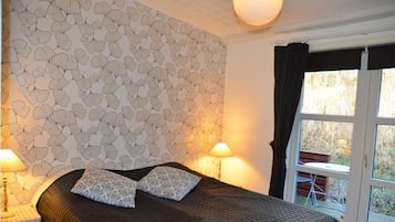 A bedroom with a patterned wallpaper, a bed with a dark quilt, two pillows, bedside lamps, and a window with curtains.