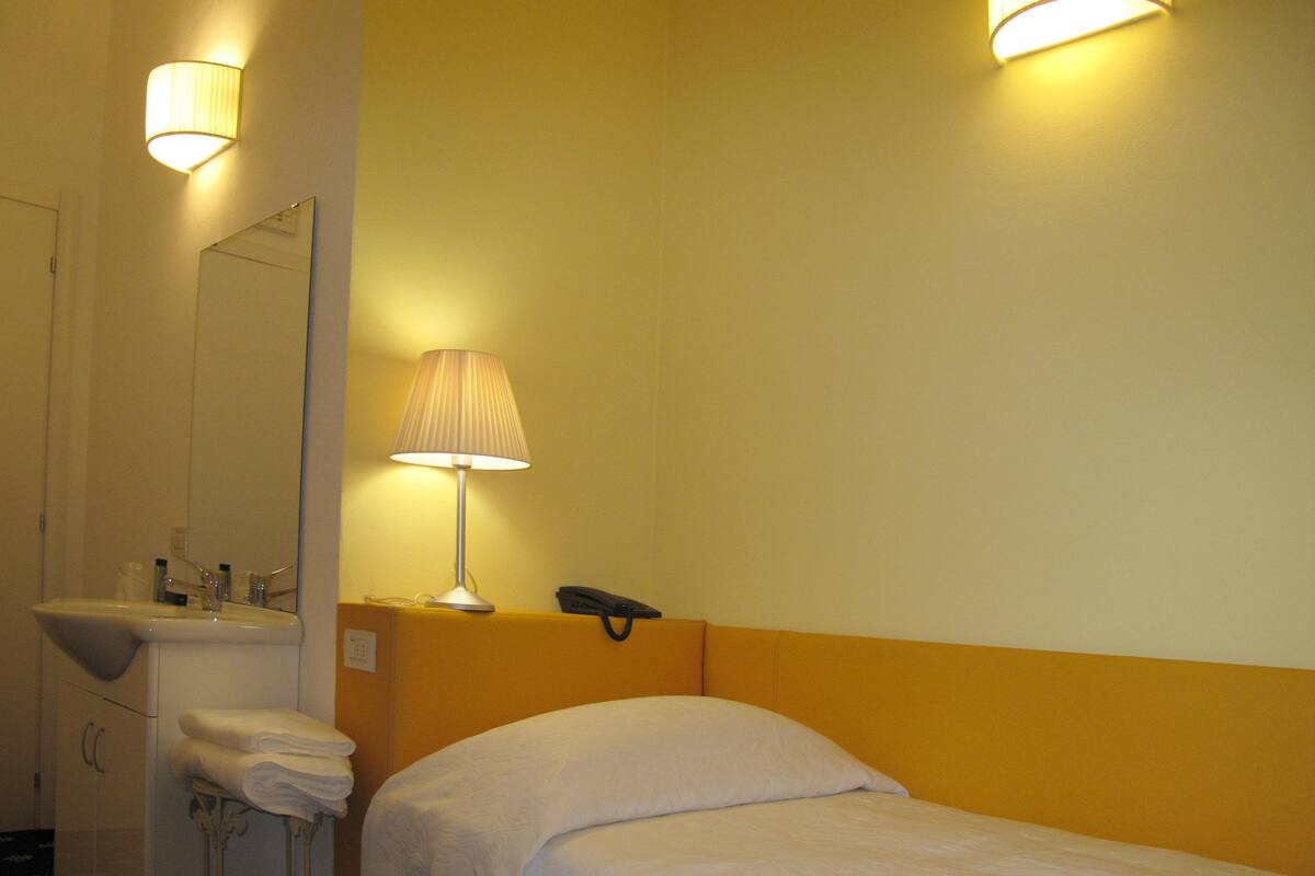 A hotel room with a bed, a bedside table, a lamp, a telephone, and a bathroom with a sink and towels.