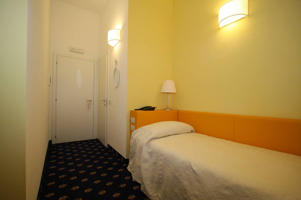 A hotel room with a single bed, a bedside table with a lamp, a phone, and a door with a handle.