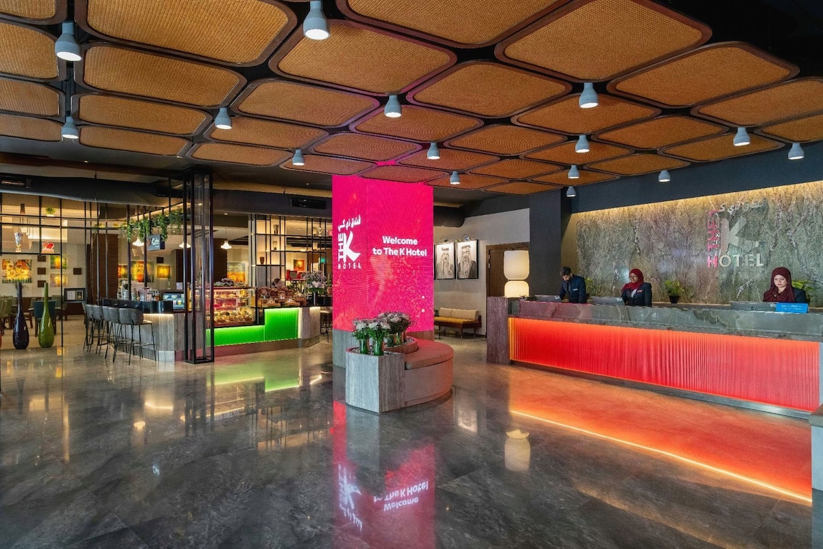 Zona de recepción de hotel con diseño moderno, que incluye un mostrador de recepción, una pantalla digital de bienvenida y un área de descanso con una pared verde.