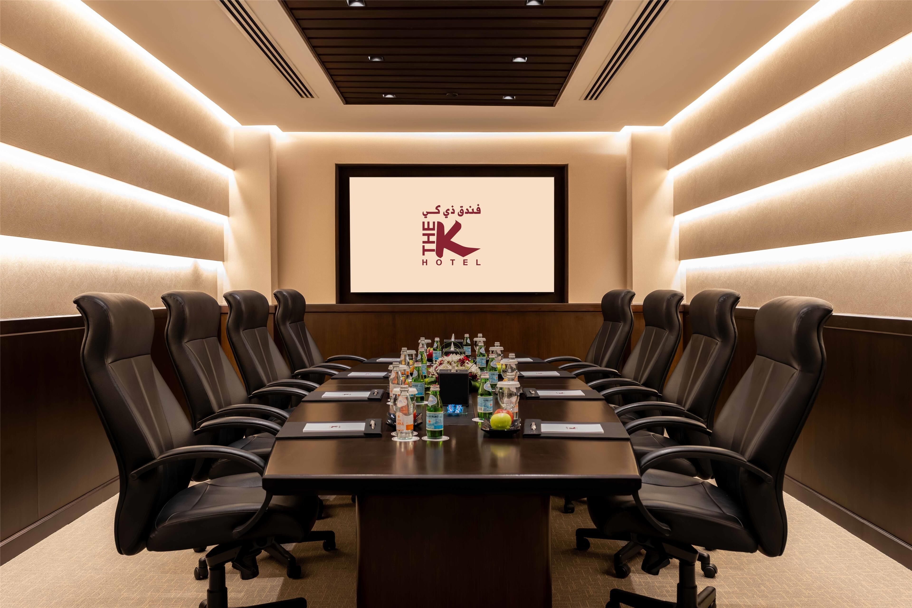 Uma sala de conferências com uma mesa comprida, várias cadeiras e uma tela de projeção exibindo "THE K HOTEL" em árabe e inglês.