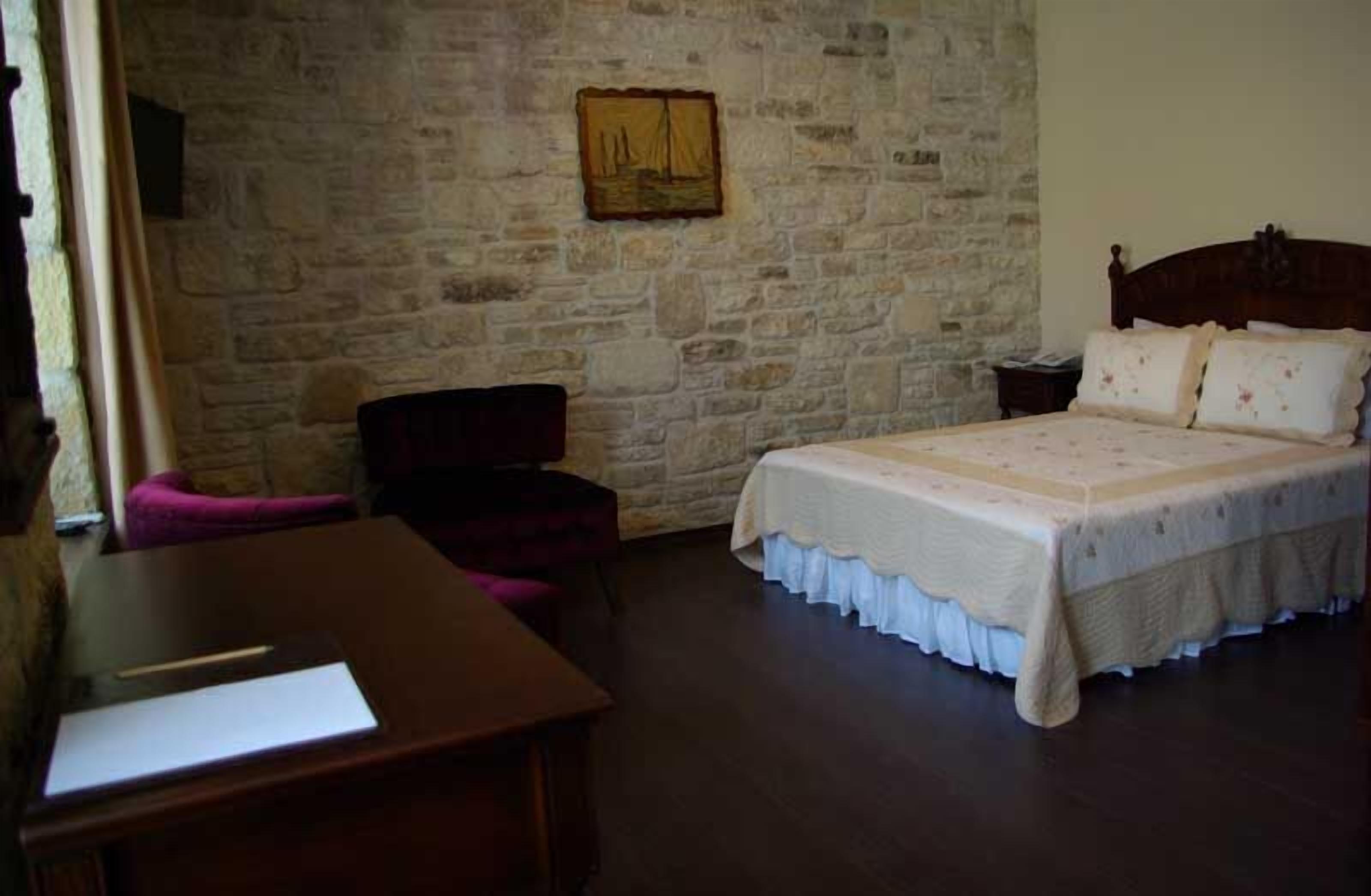 A bedroom with a stone wall, a bed with a white bedspread, a wooden nightstand, and a desk with a chair.