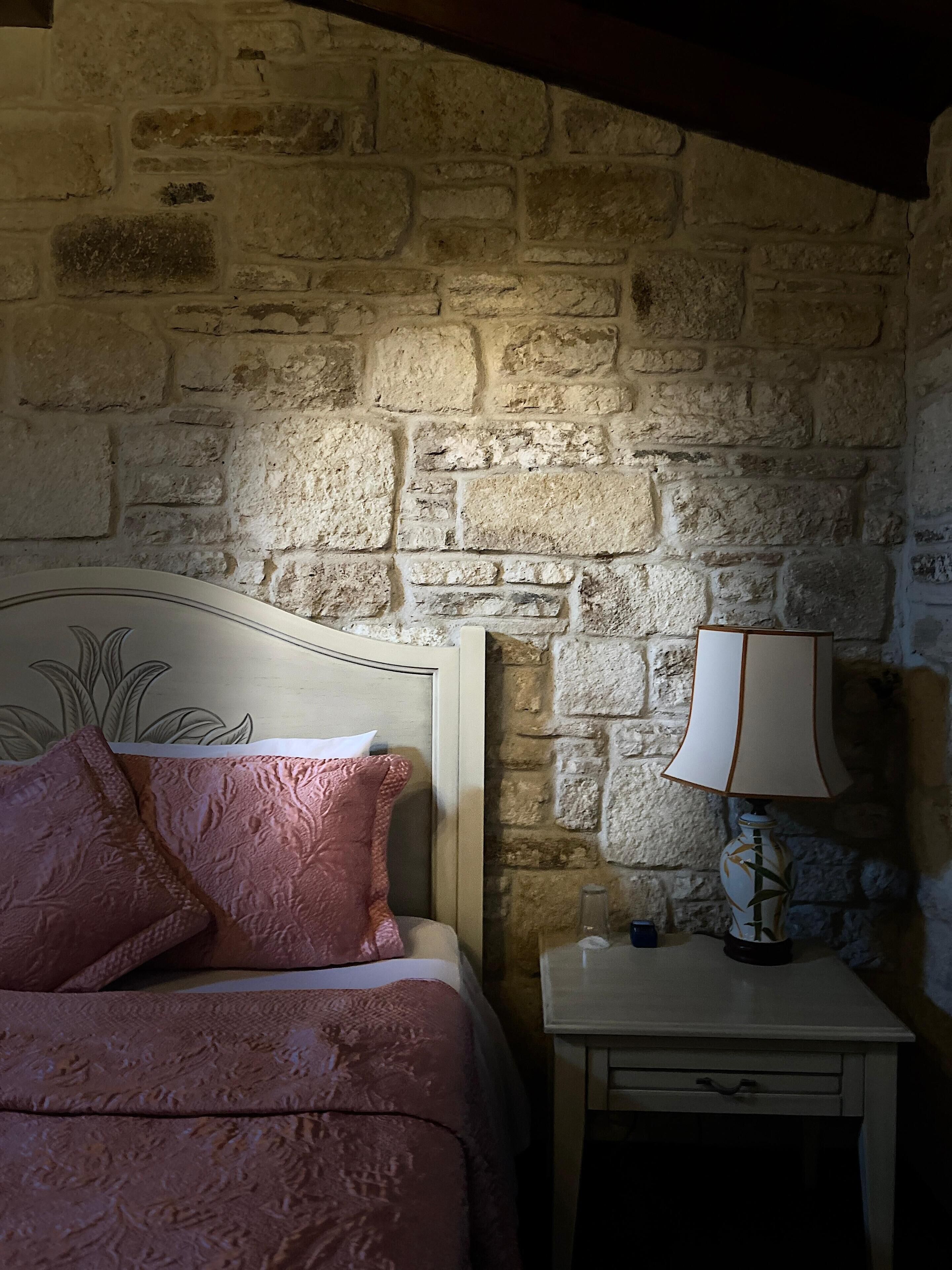 A bedroom with a stone wall, a bed with pink bedding, a white bedside table with a lamp, and a small drawer.