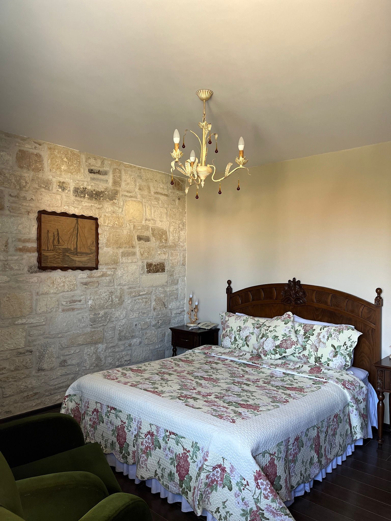 A bedroom with a stone wall, a wooden bed with floral bedding, a chandelier, and a nightstand with a lamp.