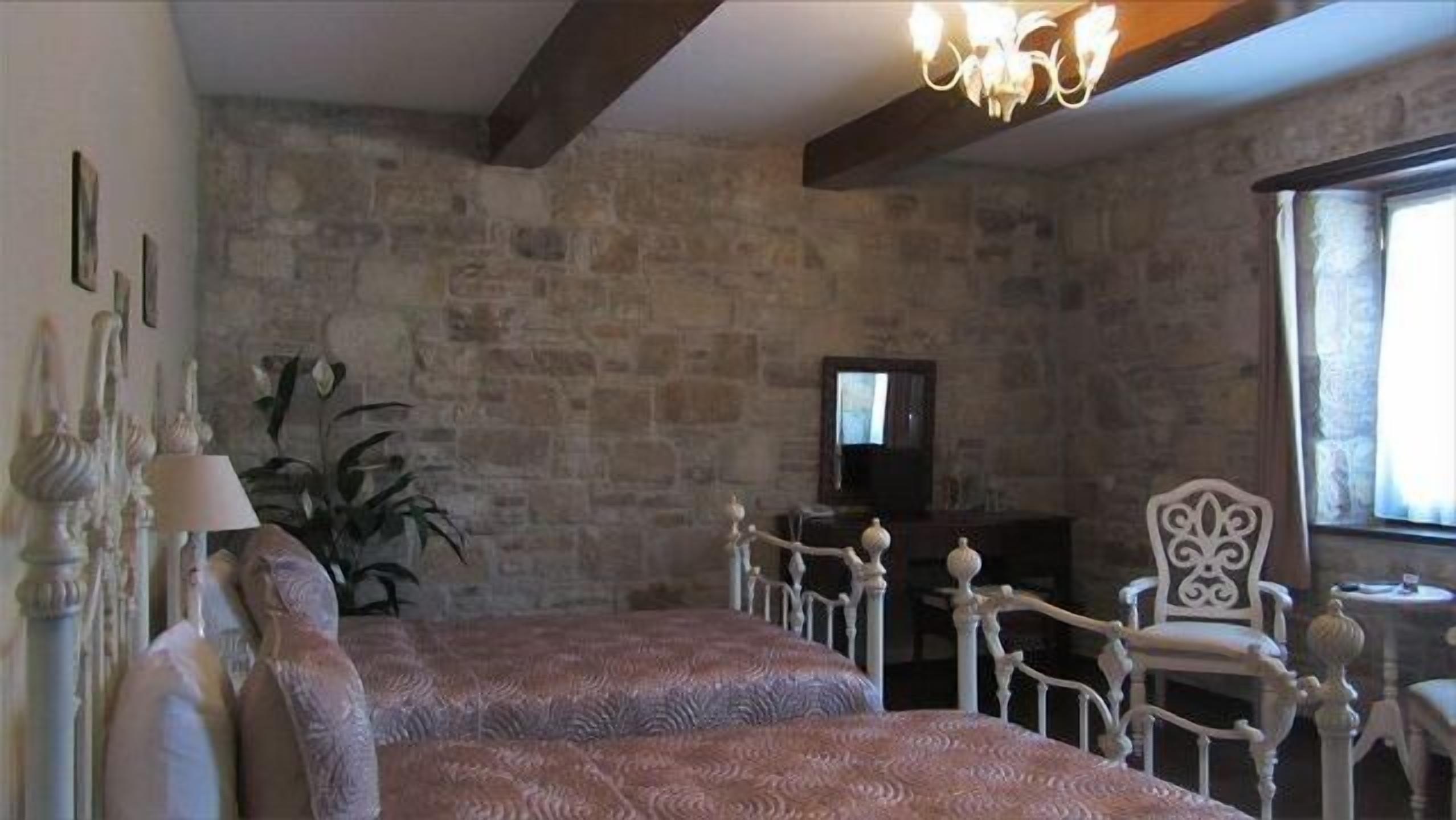 Un dormitorio con pared de piedra, una cama con colcha estampada, una silla blanca y una araña.