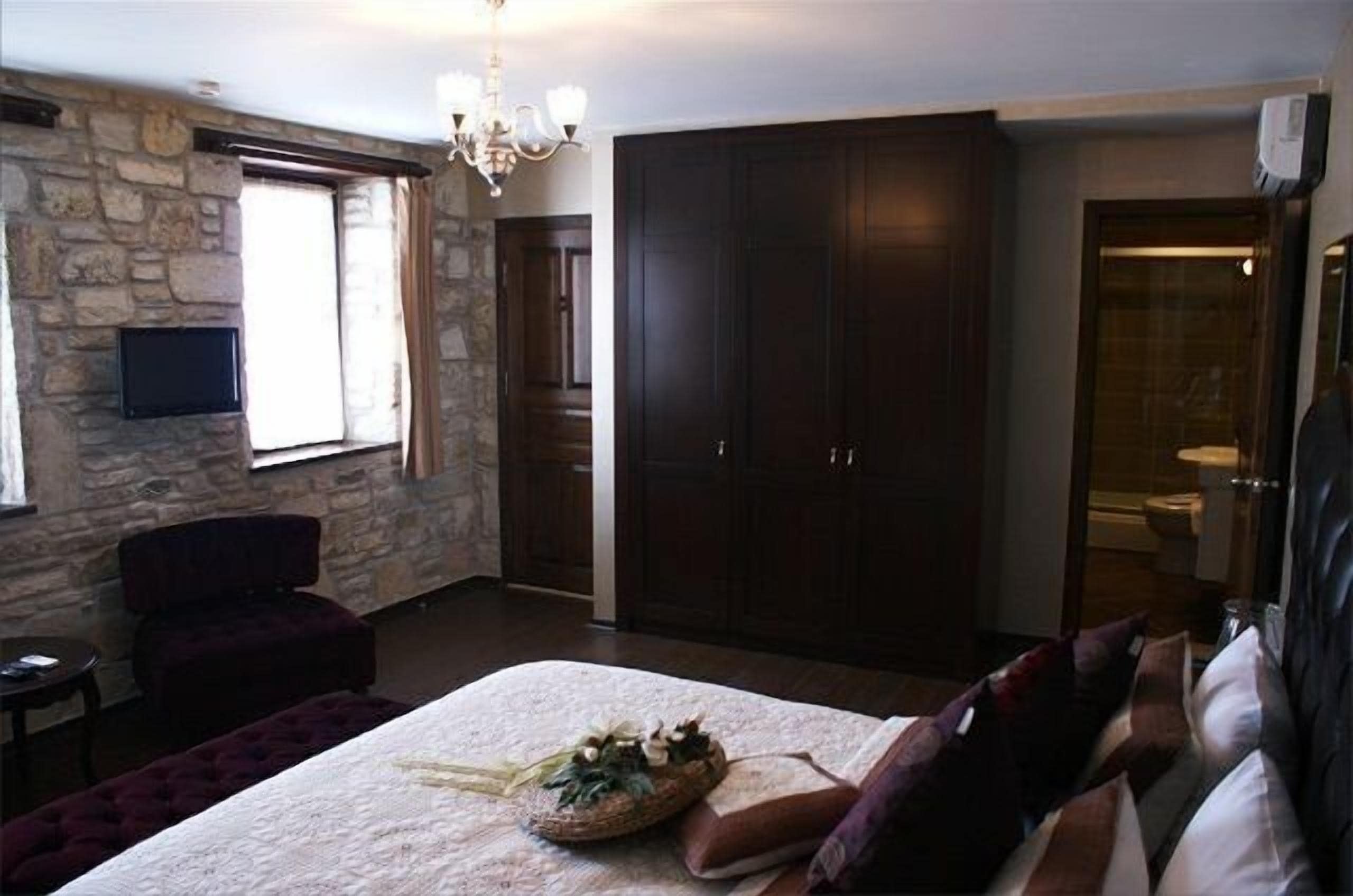 A bedroom with a stone wall, a large bed with a decorative headboard, a dark wardrobe, and a television mounted on the wall.