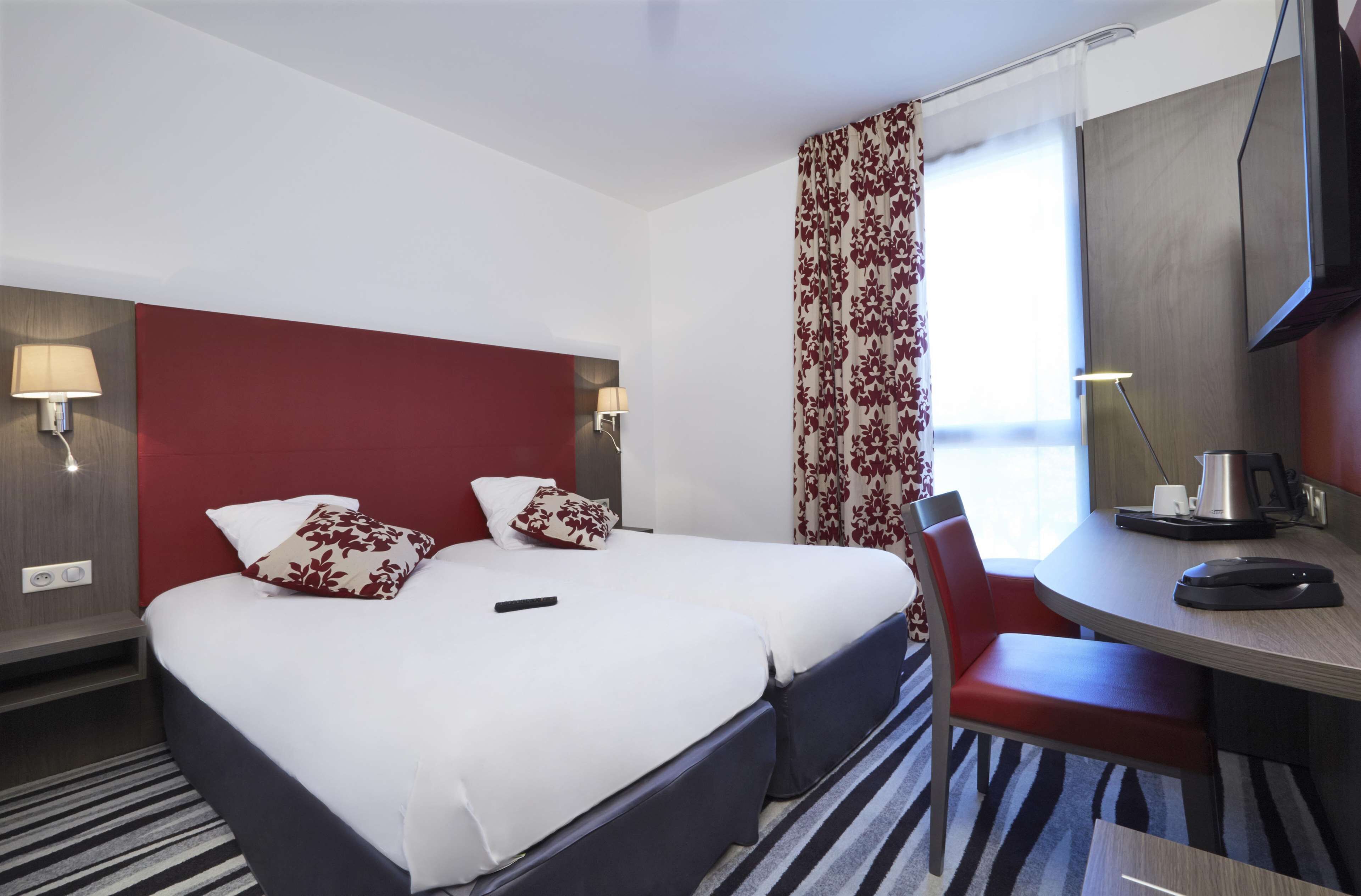 A hotel room with a double bed, a desk with a chair, a TV, and a window with curtains.