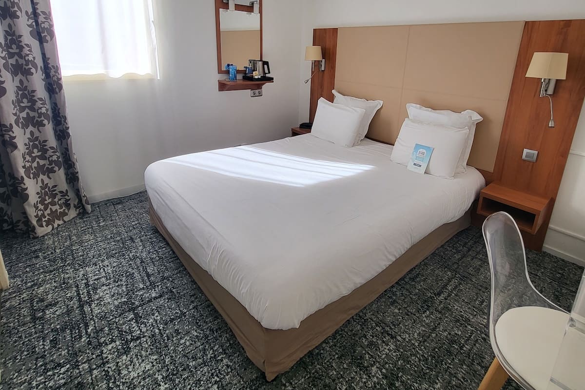 Ein Hotelzimmer mit einem großen Bett, einem hölzernen Kopfteil, einem Nachttisch mit Lampe, einer kleinen Ablage für Toilettenartikel und einem Sessel.