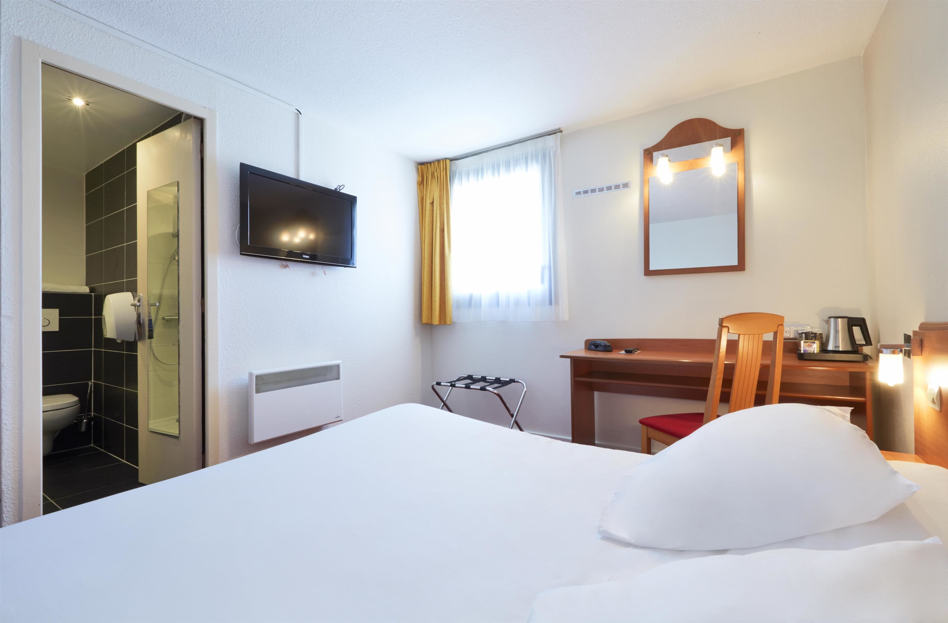 Hotellihuone, jossa on sänky, työpöytä, tuoli, televisio, peili ja ikkuna, jossa on verhot.