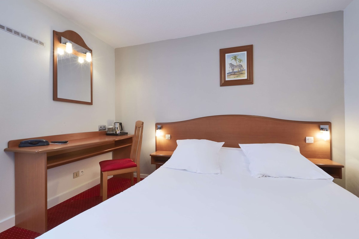 Habitación de hotel con cama, escritorio, silla, espejo y un cuadro en la pared.